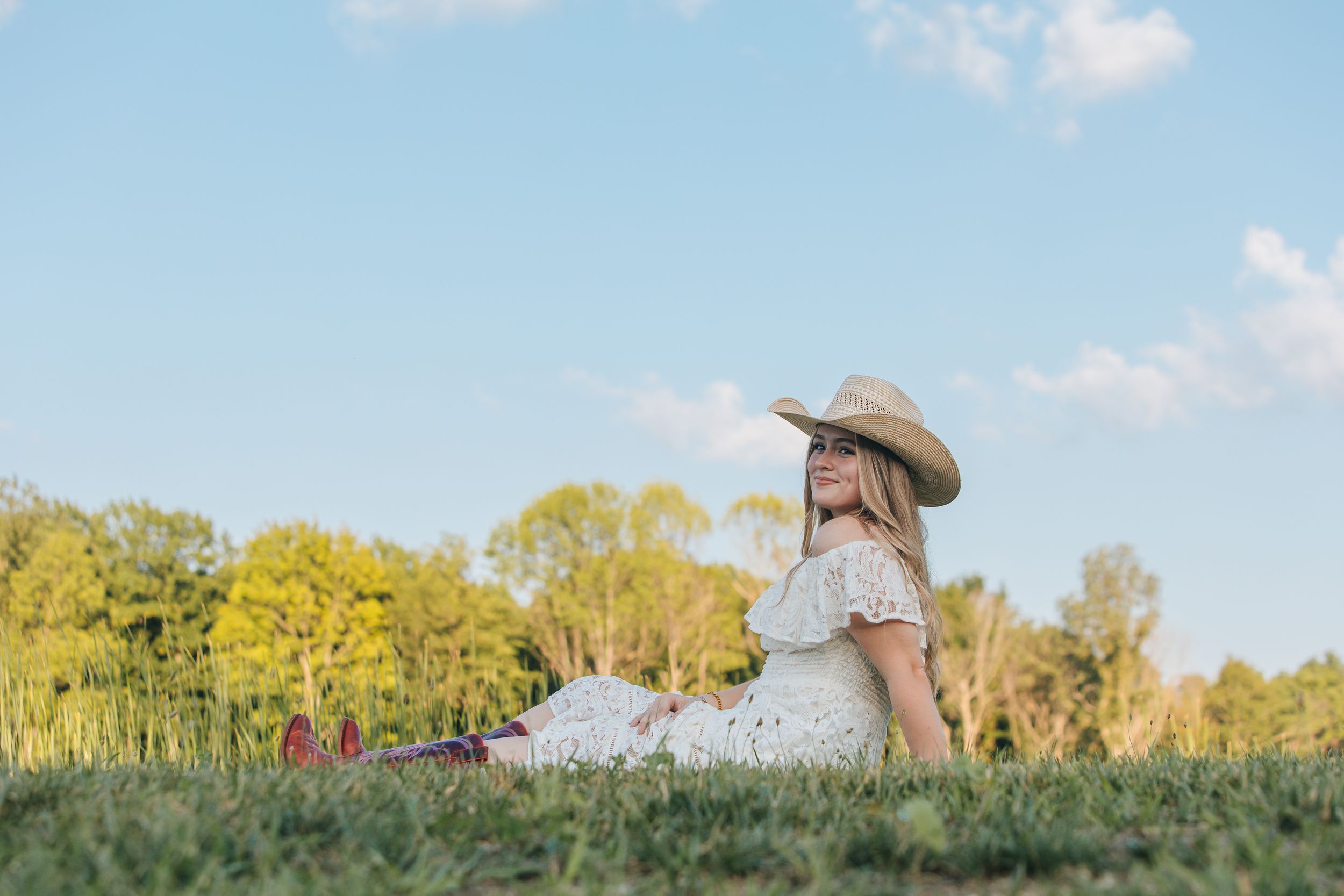 A cowgirlgirl in a white dress and cowboy hat sits on a grassy hill during a sunny summer day.