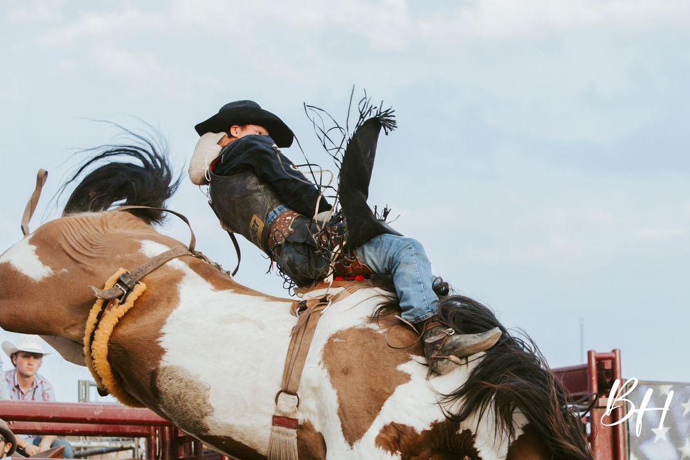 A rodeo cowboy rides a bucking bronco at a competitive rodeo event with blue sky in the background.