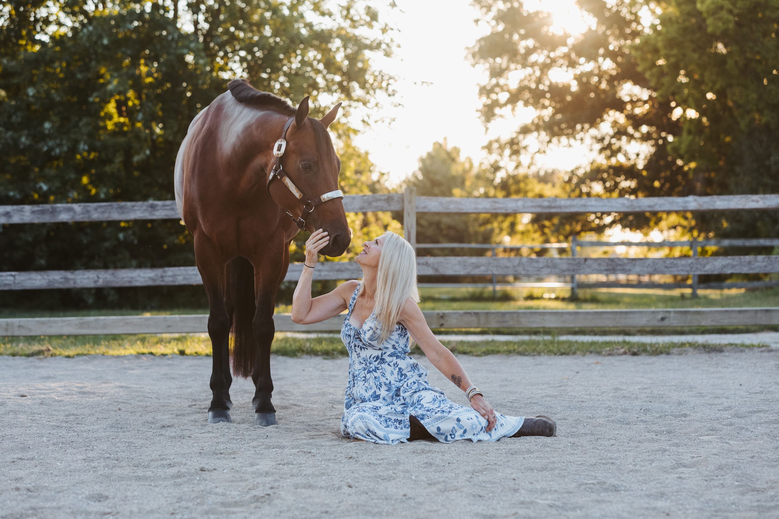 A horse and woman share a moment in a sandy arena during golden hour.