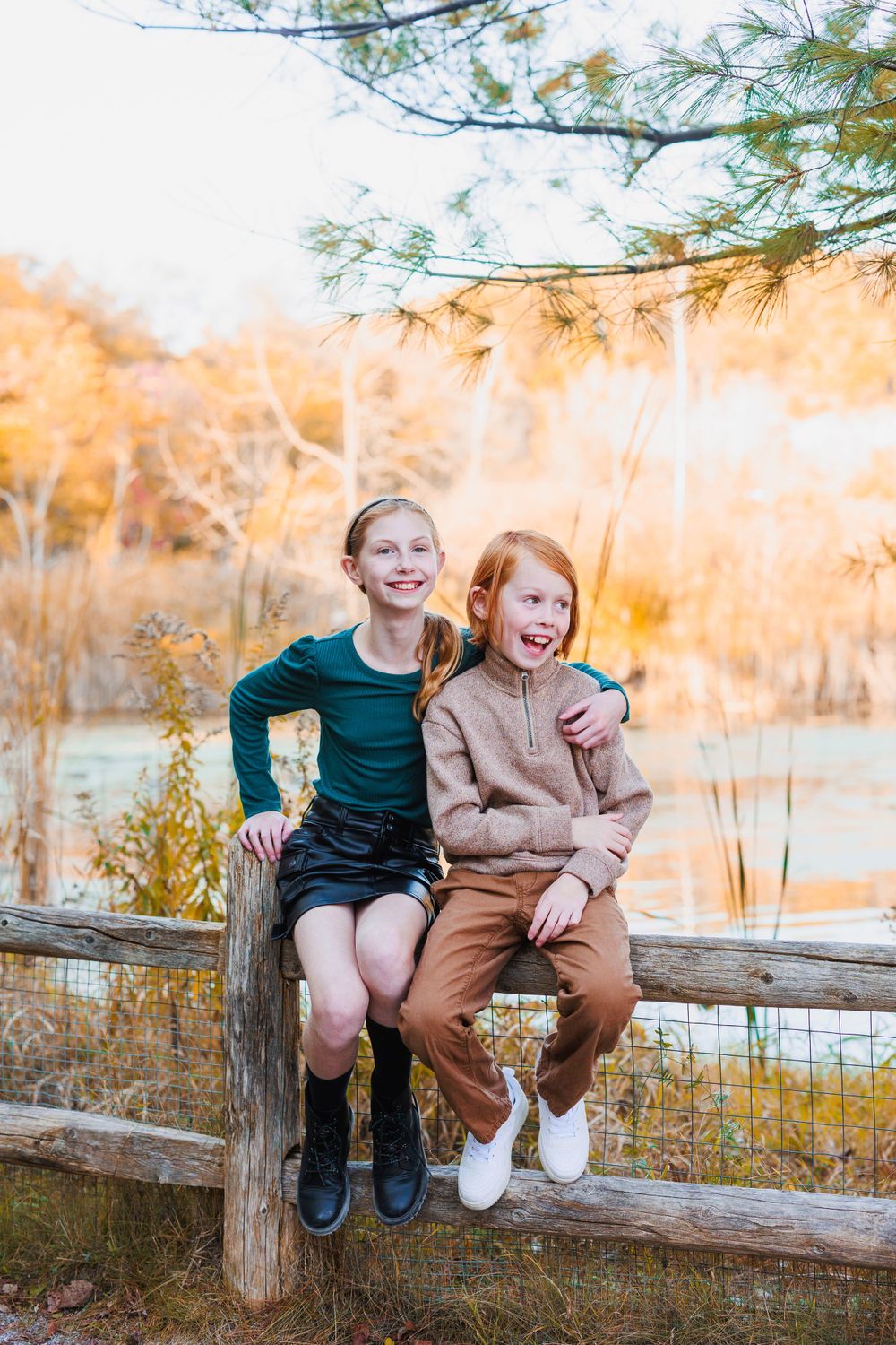 Two girls sitting on a wooden fence in a scenic autumn setting, smiling and embracing each other. Toronto family photography.