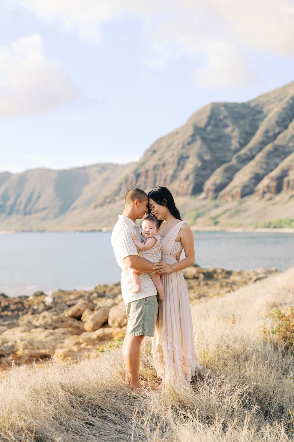 A couple holding their baby near a rocky shoreline with lush green mountains and blue water in the background.