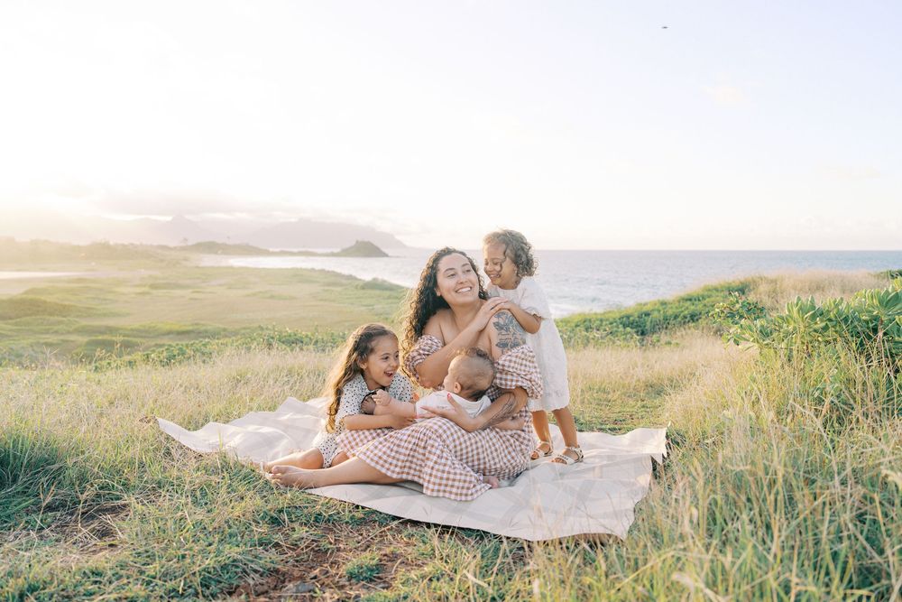 A mother sits on a blanket in a grassy coastal field, hugging her three young children in warm golden sunlight.