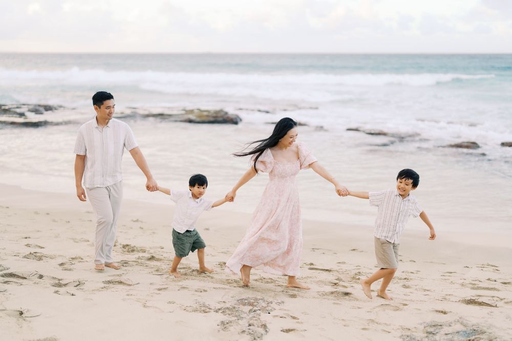 Family of four walking hand-in-hand on a sandy beach, parents and two young boys enjoying a sunny day by the ocean.
