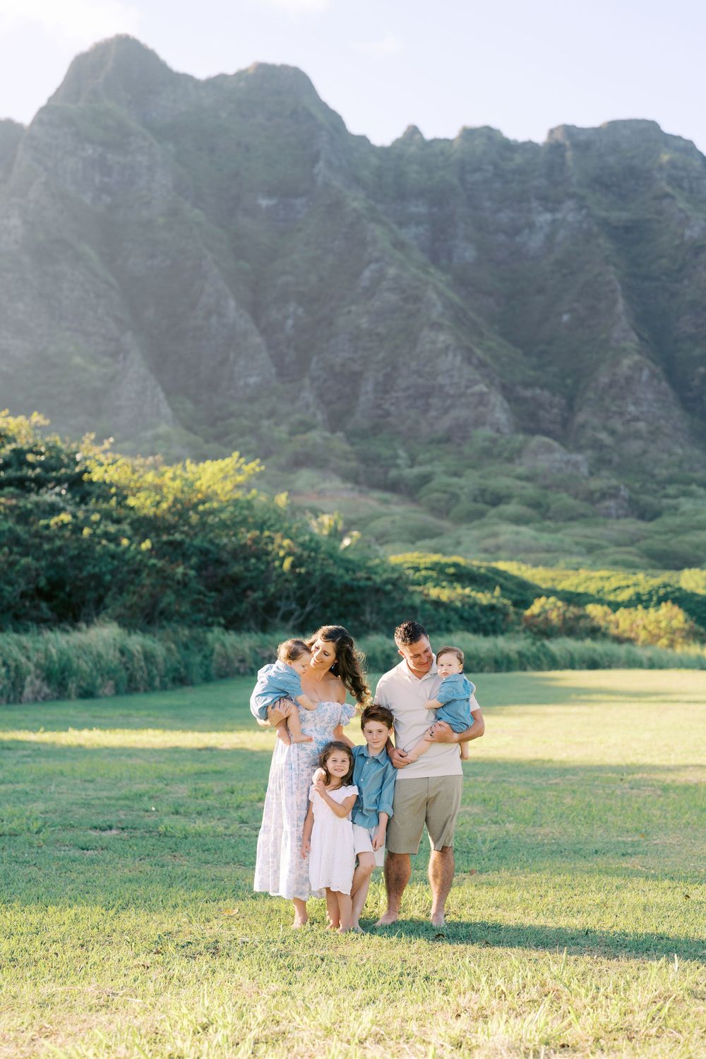 Family of five poses on green lawn with lush mountain backdrop in Hawaii, parents holding babies with young girl between them.