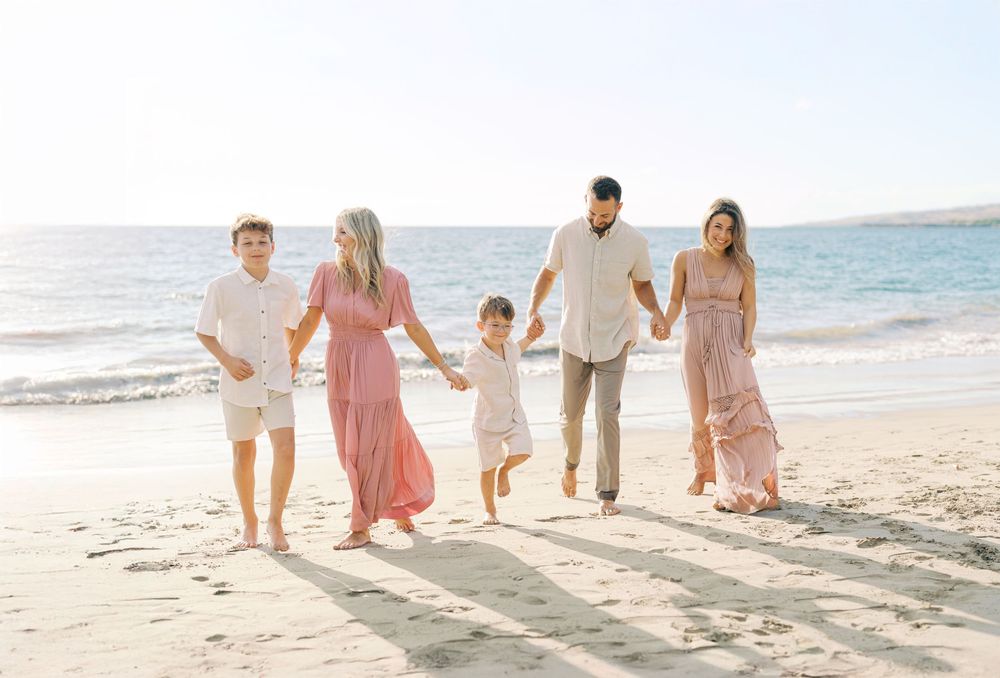 Family of five walking hand-in-hand on a sunny beach, dressed in coordinating pink and white outfits.