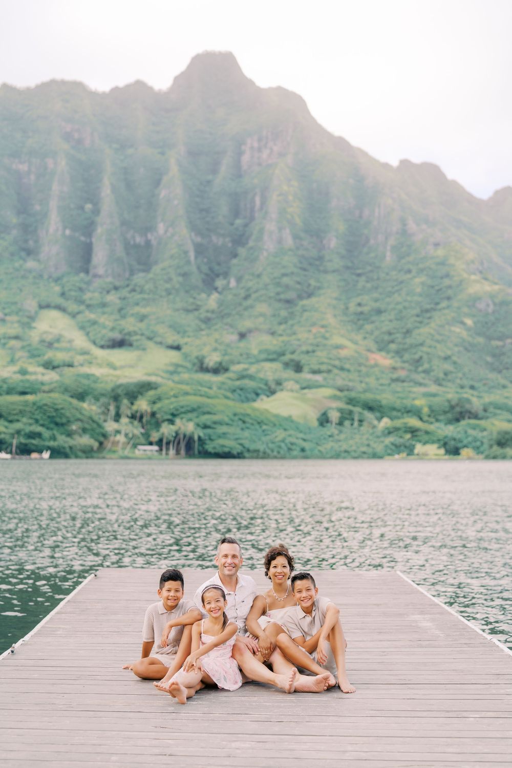 Family of five sitting on a dock with lush green mountains and calm water in Hawaii.