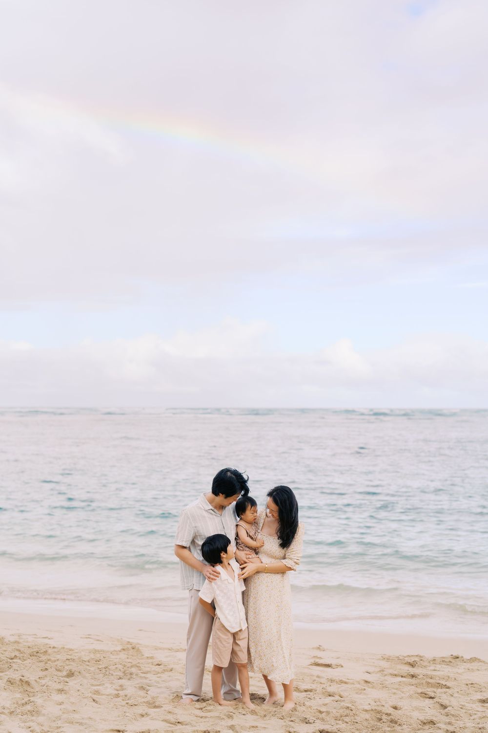 Family of four in neutral tones embracing on a sandy beach with ocean waves and a rainbow in the background.