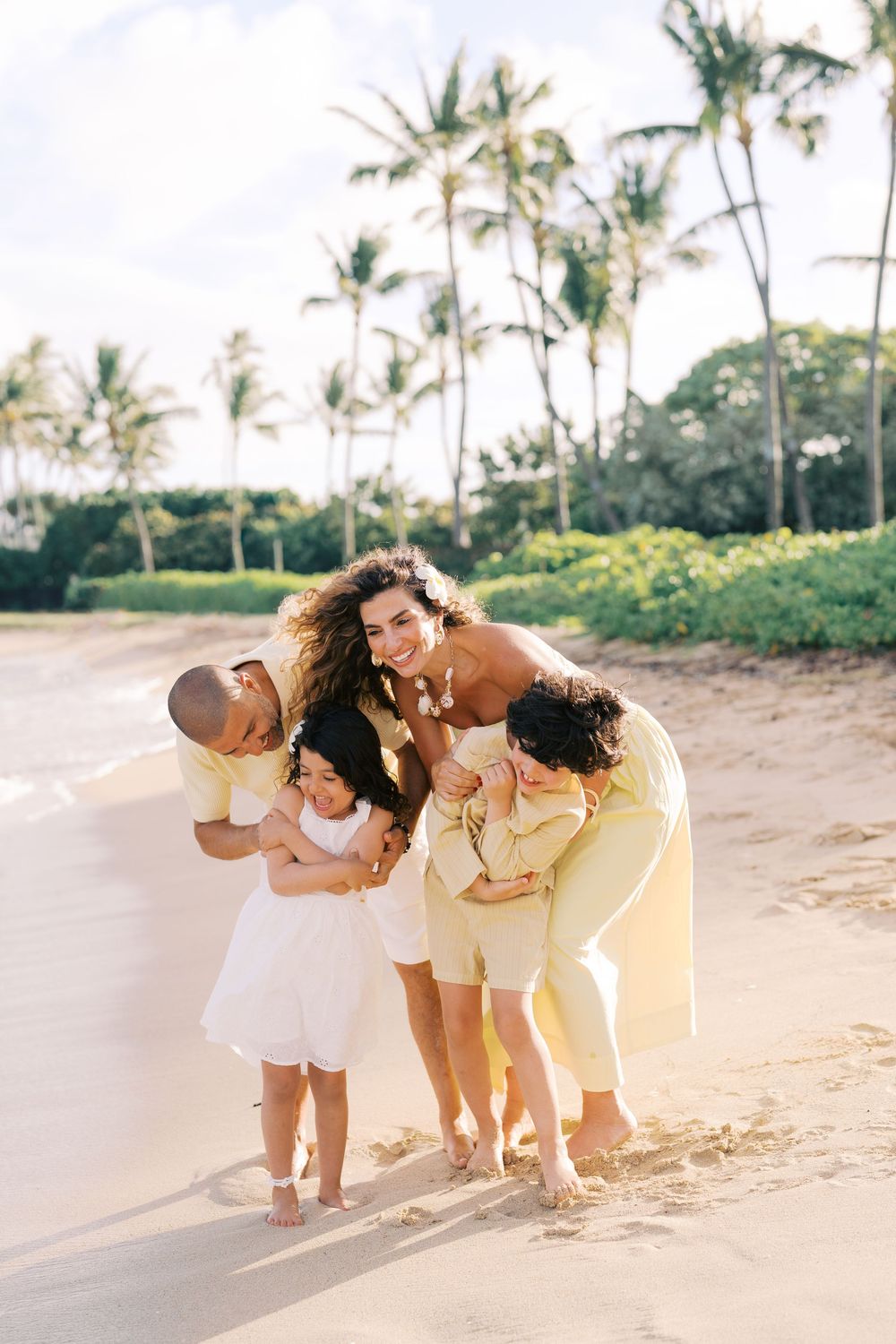Family in yellow outfits laughing together on a sunny tropical beach with palm trees in the background.