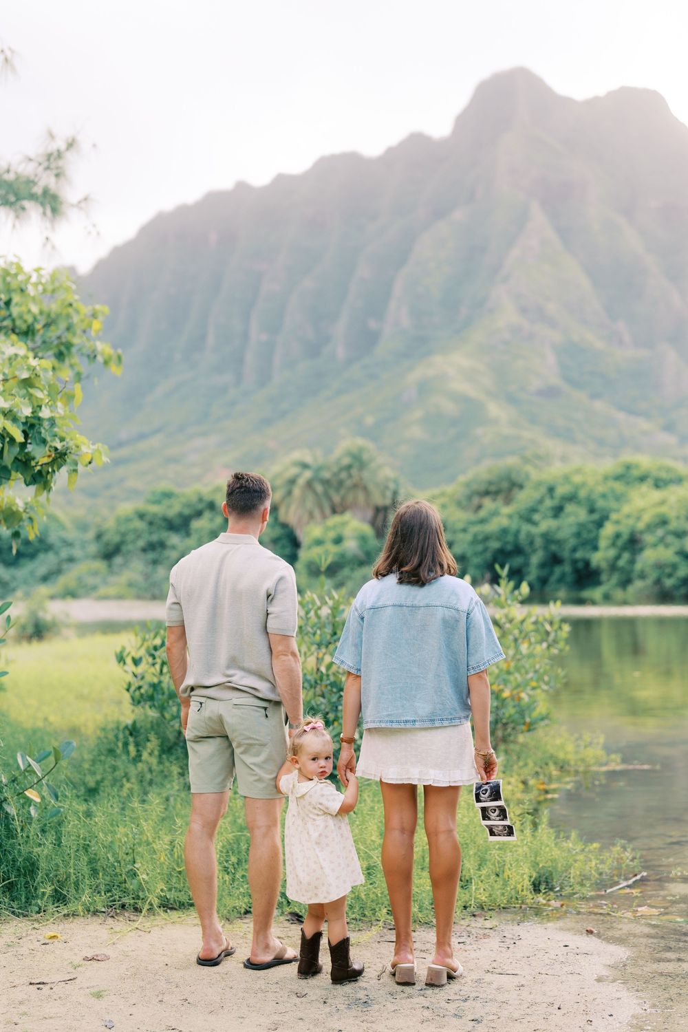 Family of three holding hands facing lush green mountains, woman holding ultrasound photo, pregnancy announcement in Hawaii.