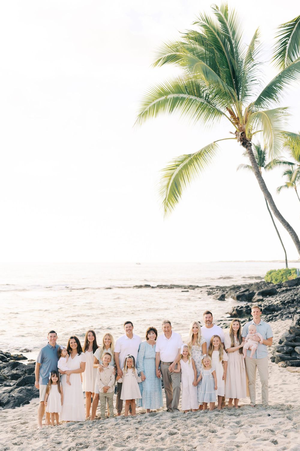 Large family group portrait on a sandy Hawaiian beach with palm trees and ocean backdrop at golden hour.