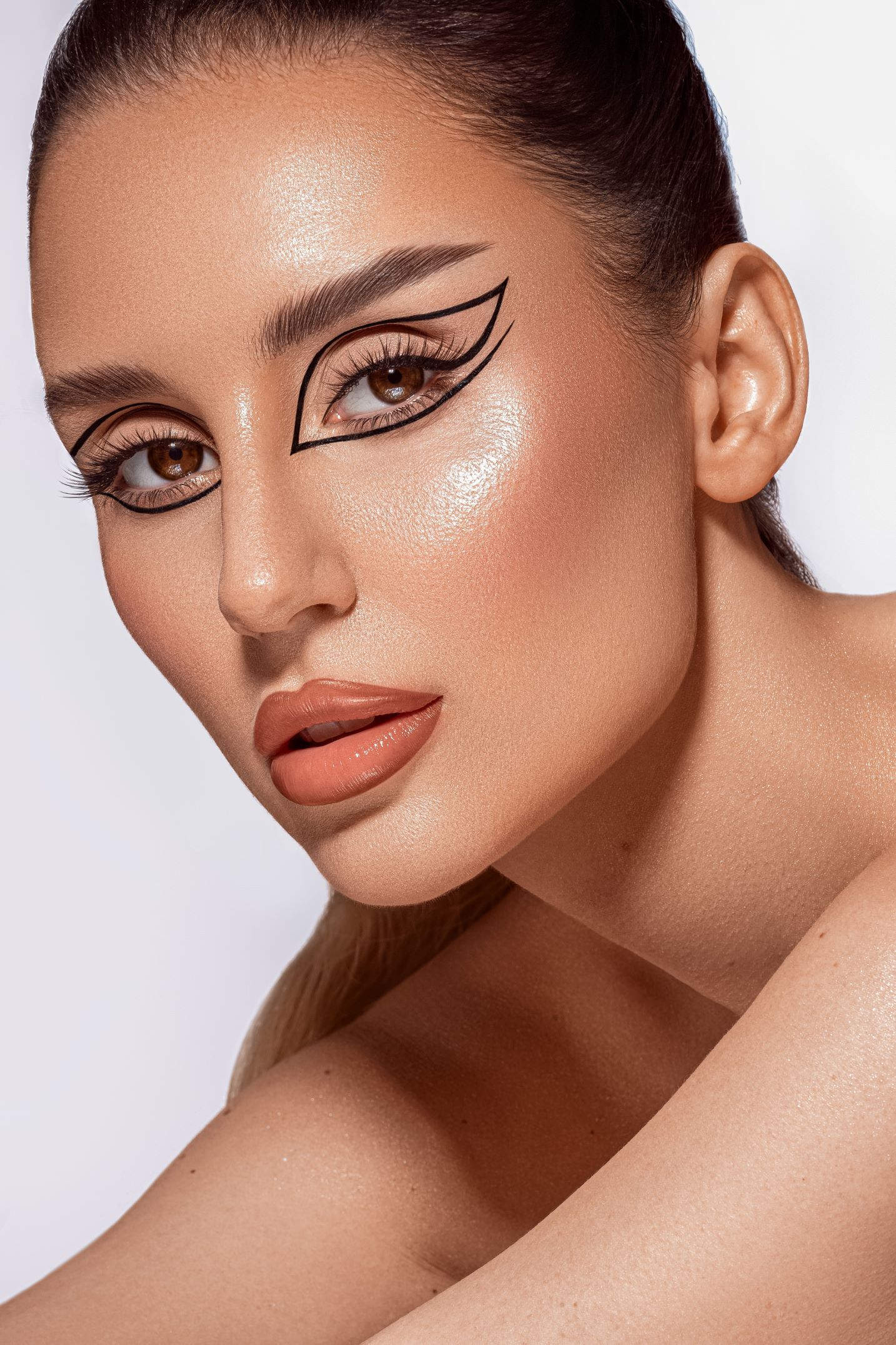 Dramatic cat eye makeup look with sharp winged liner, matte coral lipstick and glowing highlighted skin against white background.