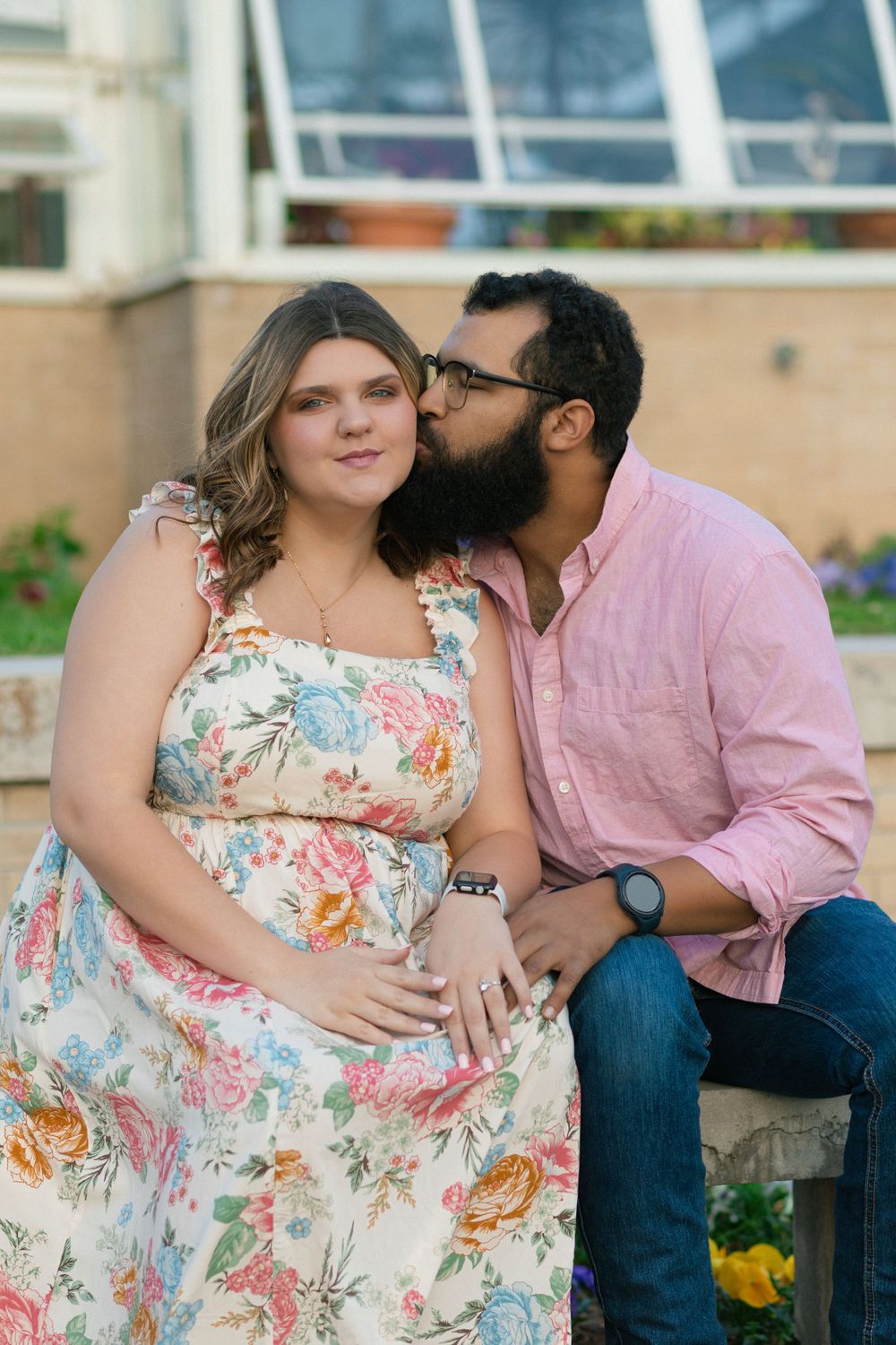 Couple in coordinating floral dress and pink shirt share an intimate moment on outdoor bench.