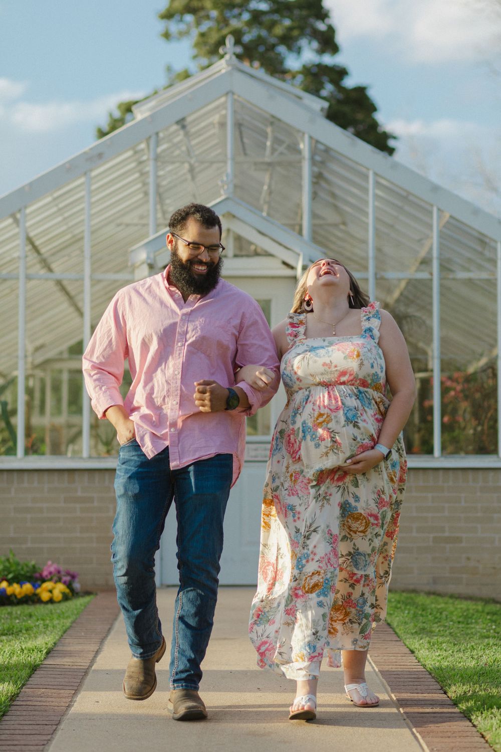 A couple walks together laughing on a pathway in front of a greenhouse surrounded by colorful flowers.