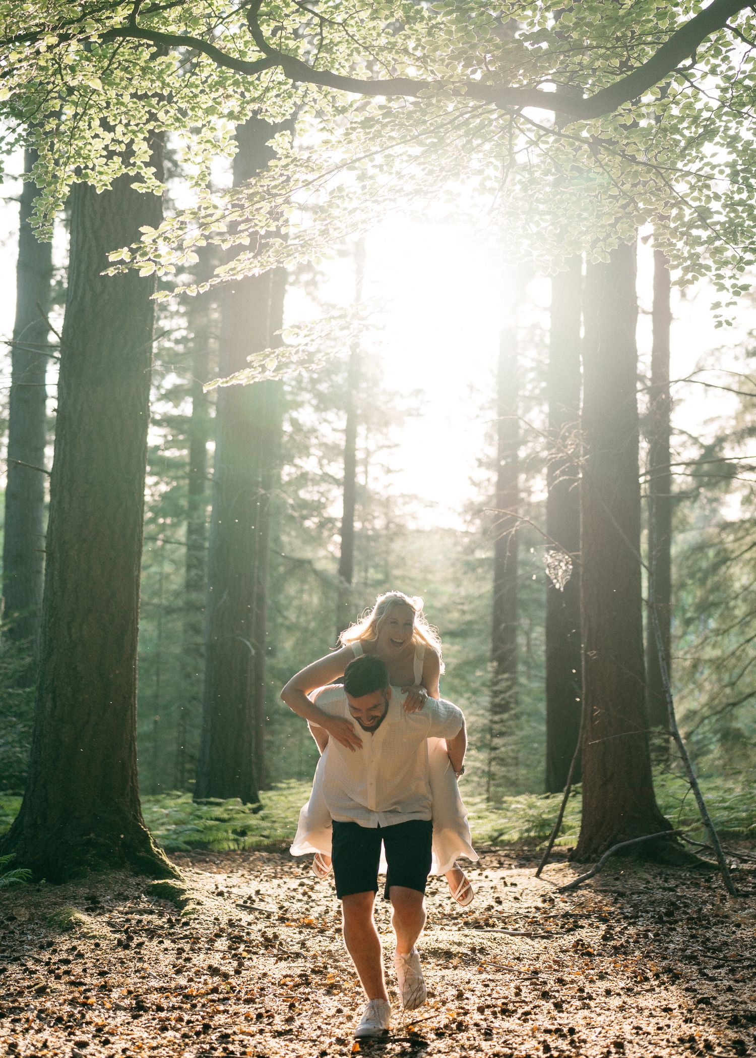 Couple sharing a piggyback ride through a sunlit forest with tall trees and golden light filtering through leaves.