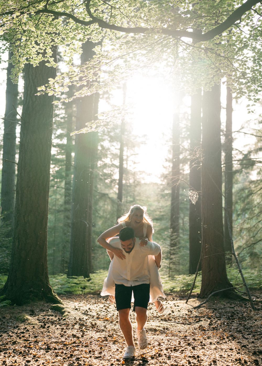 Couple sharing a piggyback ride through a sunlit forest with tall trees and golden light filtering through leaves.
