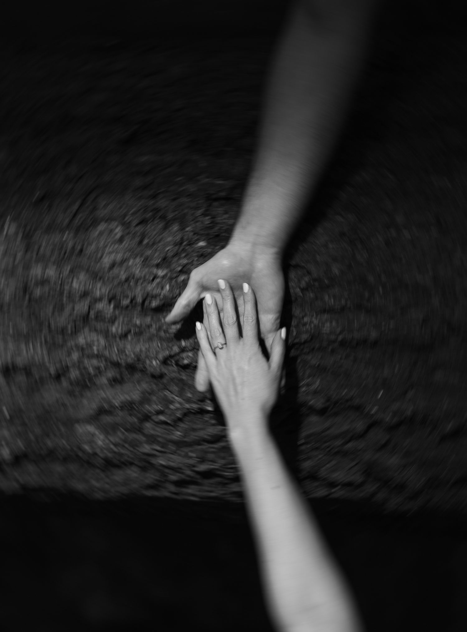 Black and white photo of two hands gently touching, one wearing a ring, over a blurred dark background.