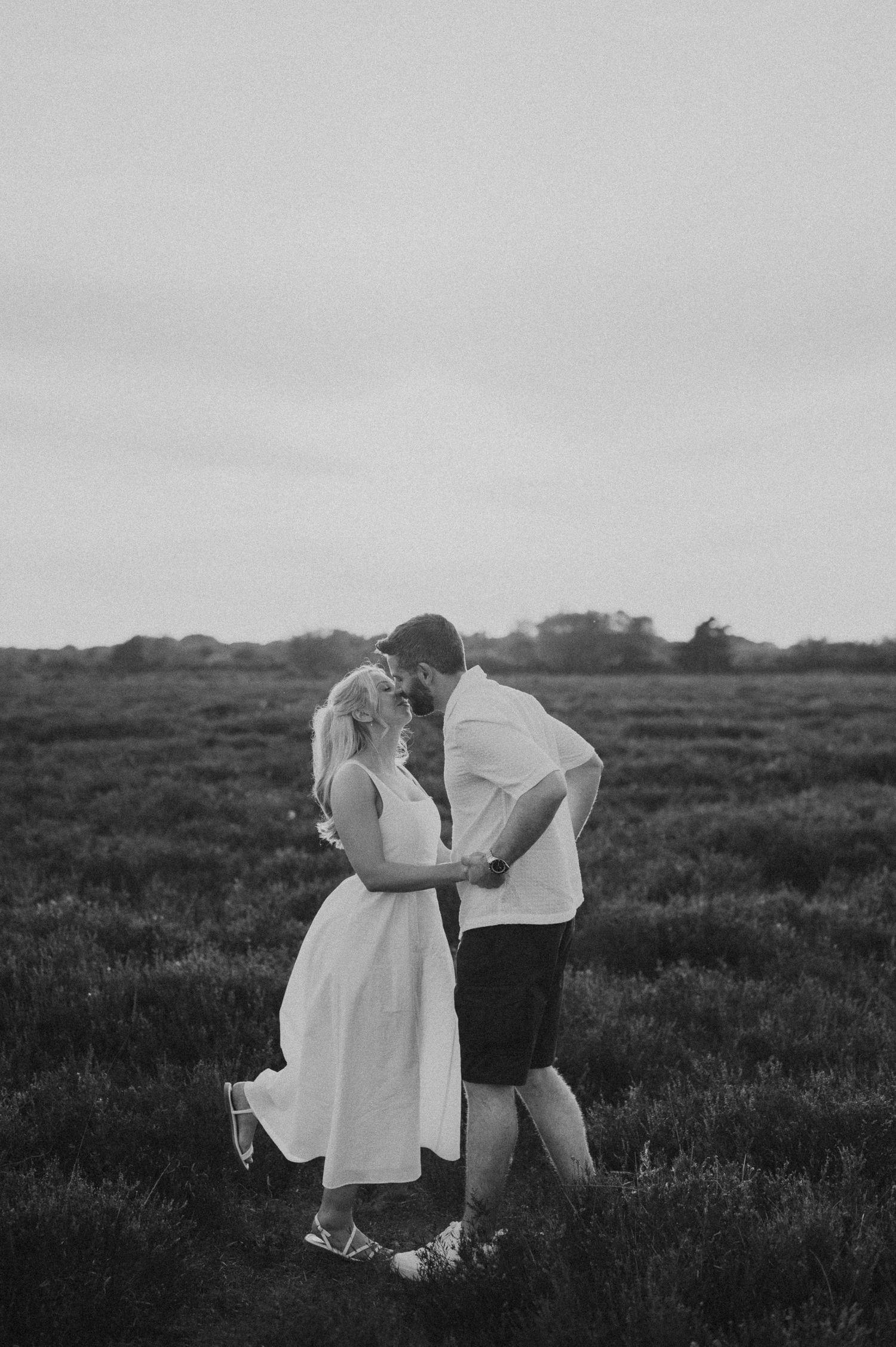 Black and white photo of a couple sharing a romantic moment in an open field at dusk.