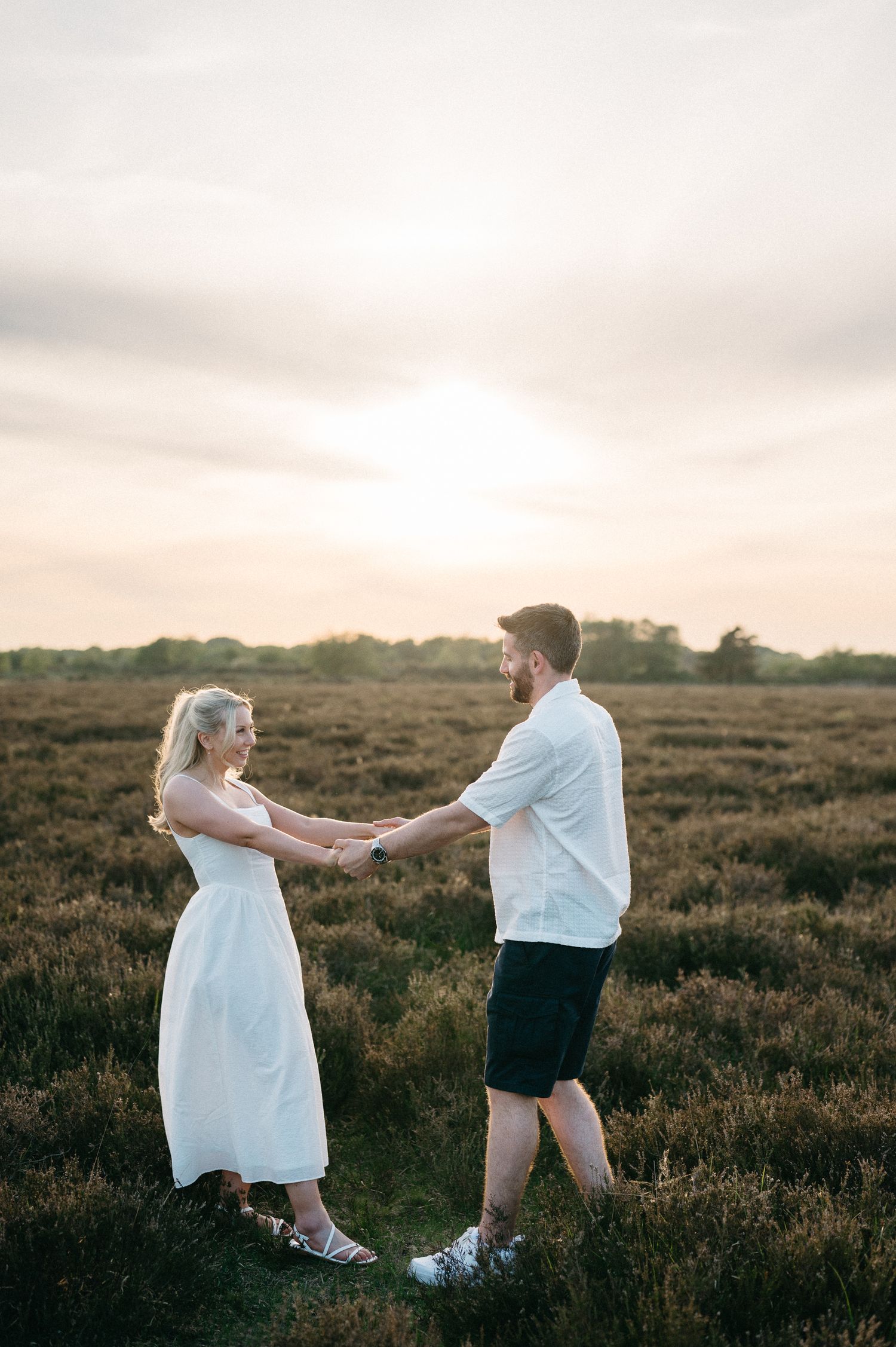 Couple holding hands in a golden field at sunset, woman in white dress, man in white shirt and black shorts.