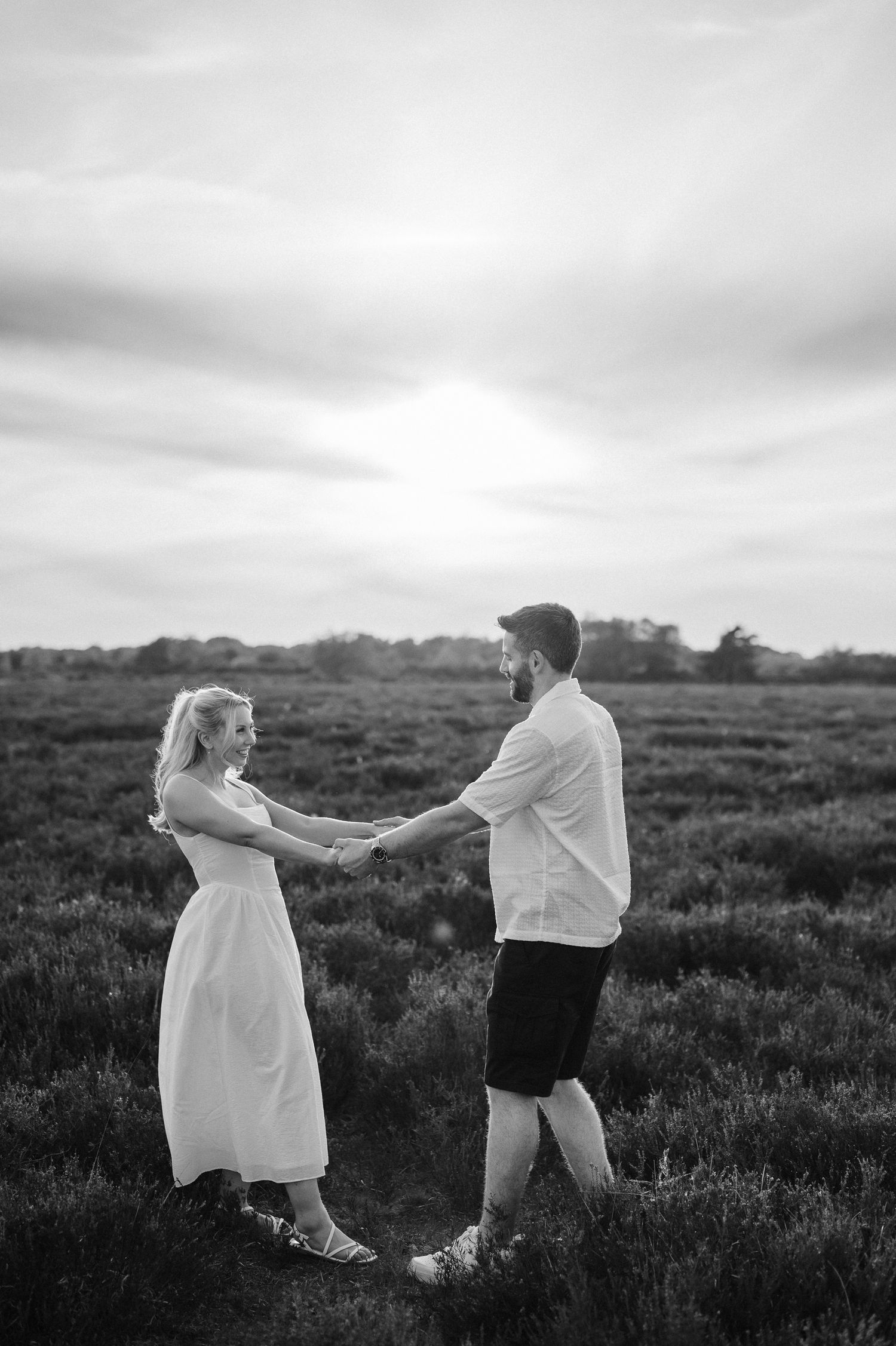 Black and white photo of a couple holding hands in a field at sunset, woman in white dress facing bearded man.