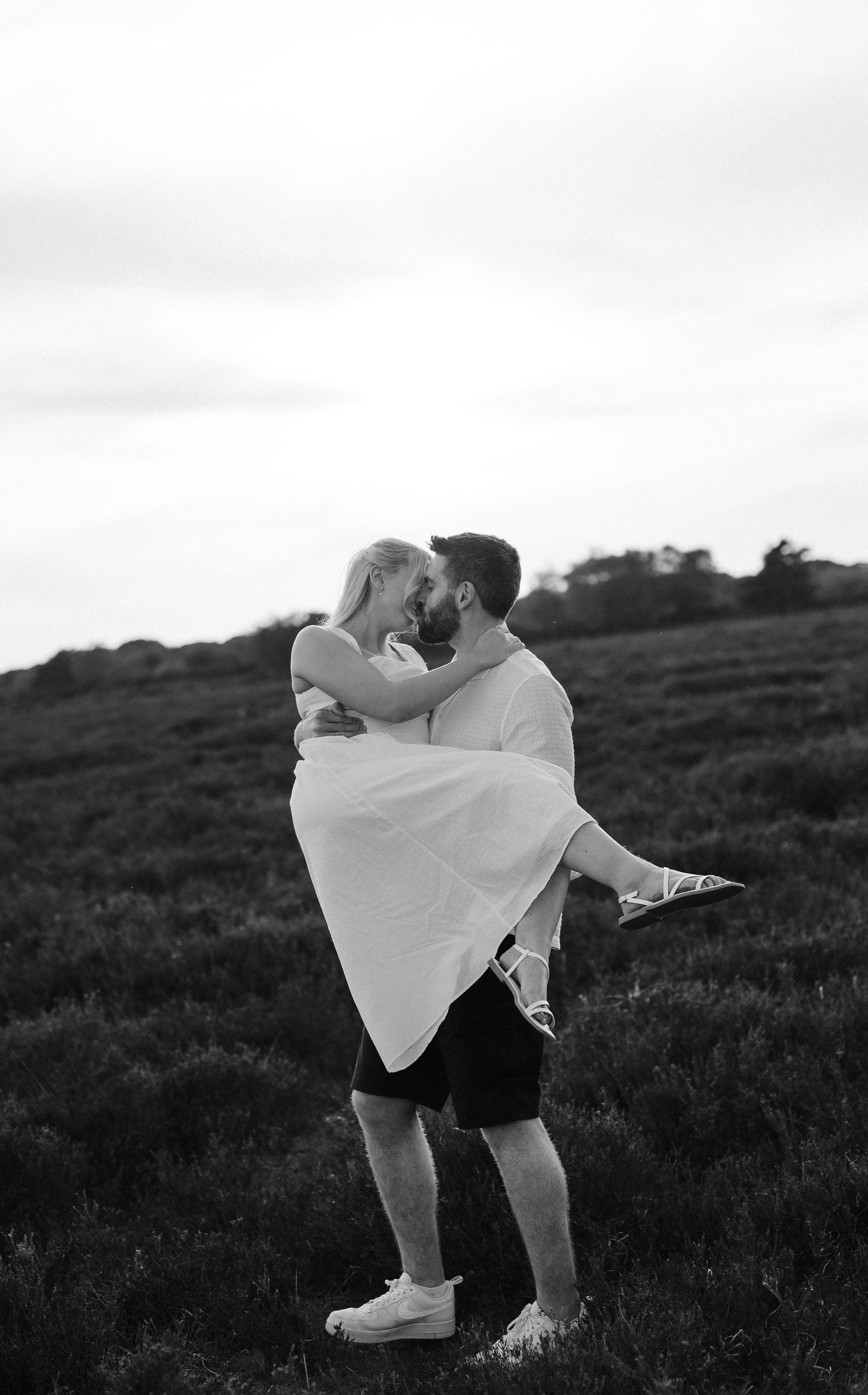 Black and white photo of a man lifting and kissing a woman in a field at dusk.