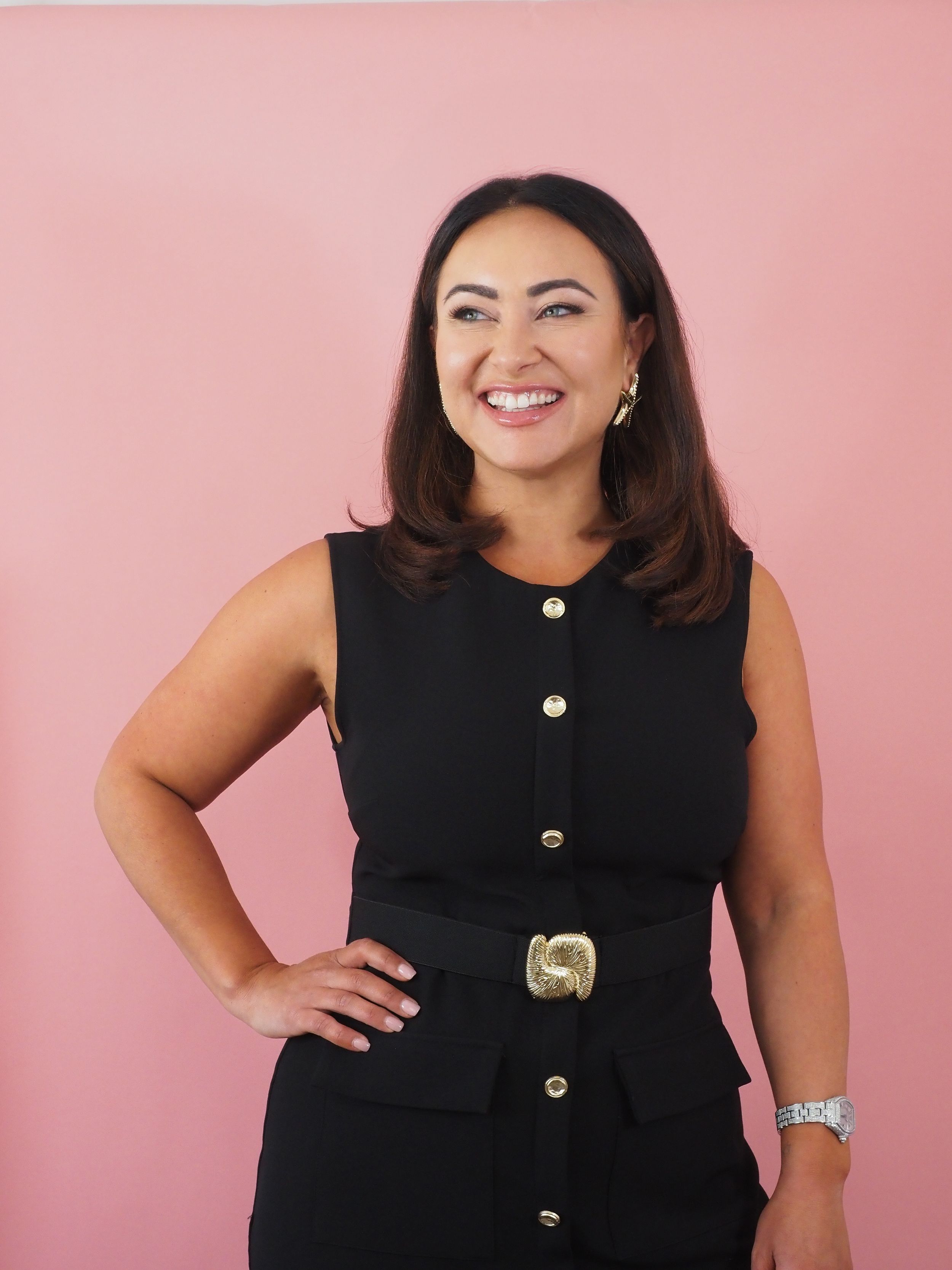 Woman in a sleek black button-up dress with gold belt against a pink backdrop.