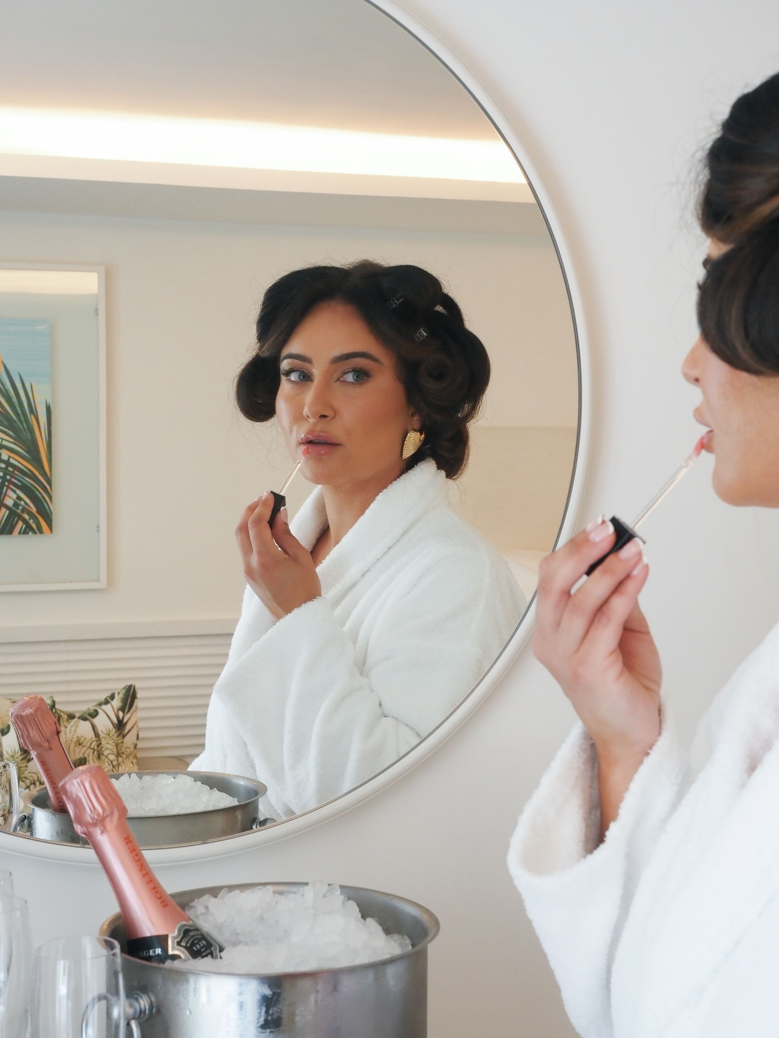 A person in a white bathrobe applies makeup in a circular bathroom mirror, creating a repeating reflection pattern.