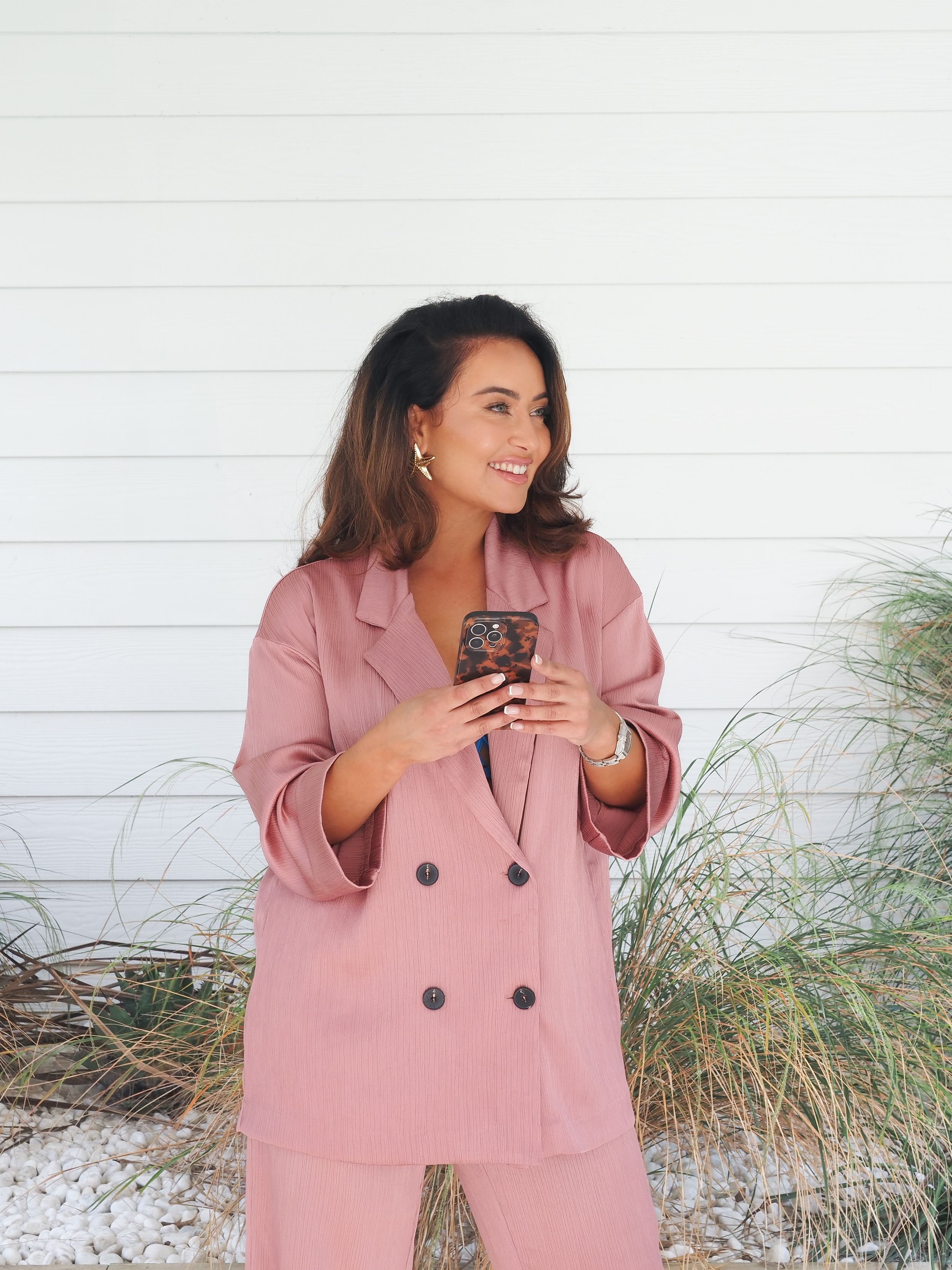 Smiling woman wearing a pink pantsuit and holding a phone against a white shiplap wall with ornamental grass.