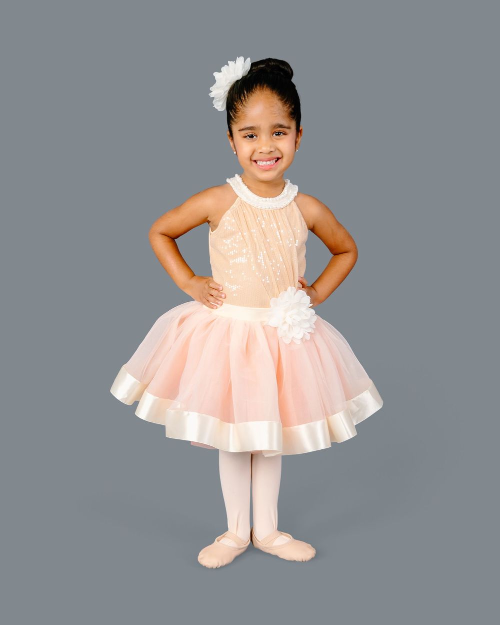 Young dancer in pink tulle dress and white tights poses gracefully against a gray studio backdrop.