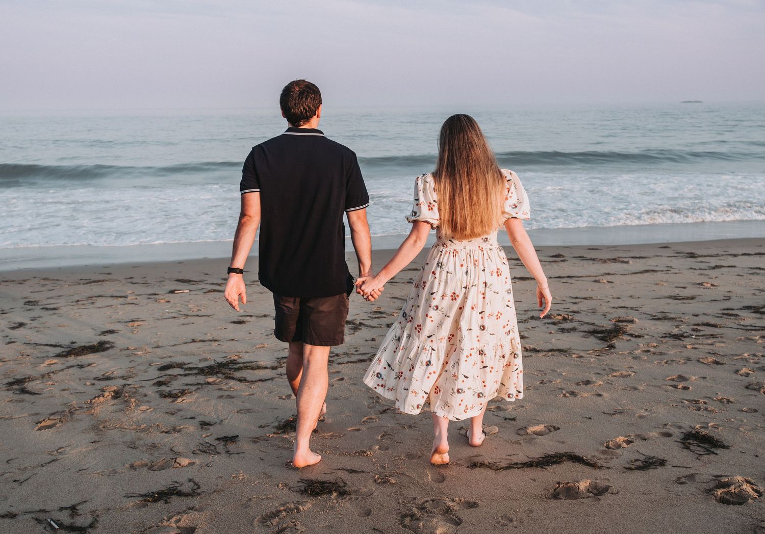 A newly engaged couple walks together hand in hand at Reid State Park at sunset by the ocean waves.