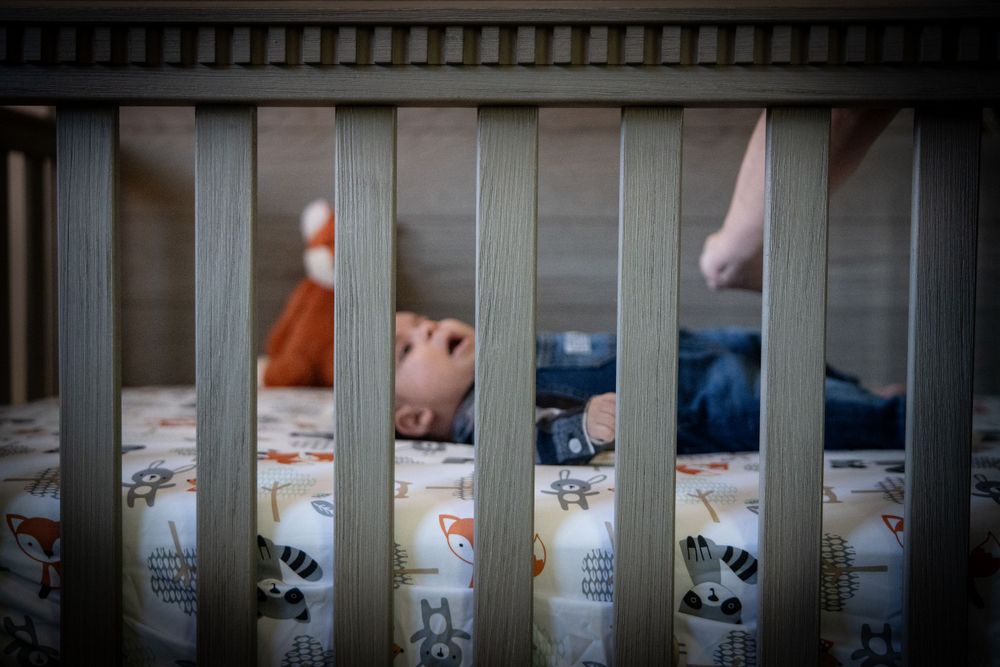 A sleeping baby in jeans and an orange shirt lies peacefully in a wooden crib with patterned bedding.