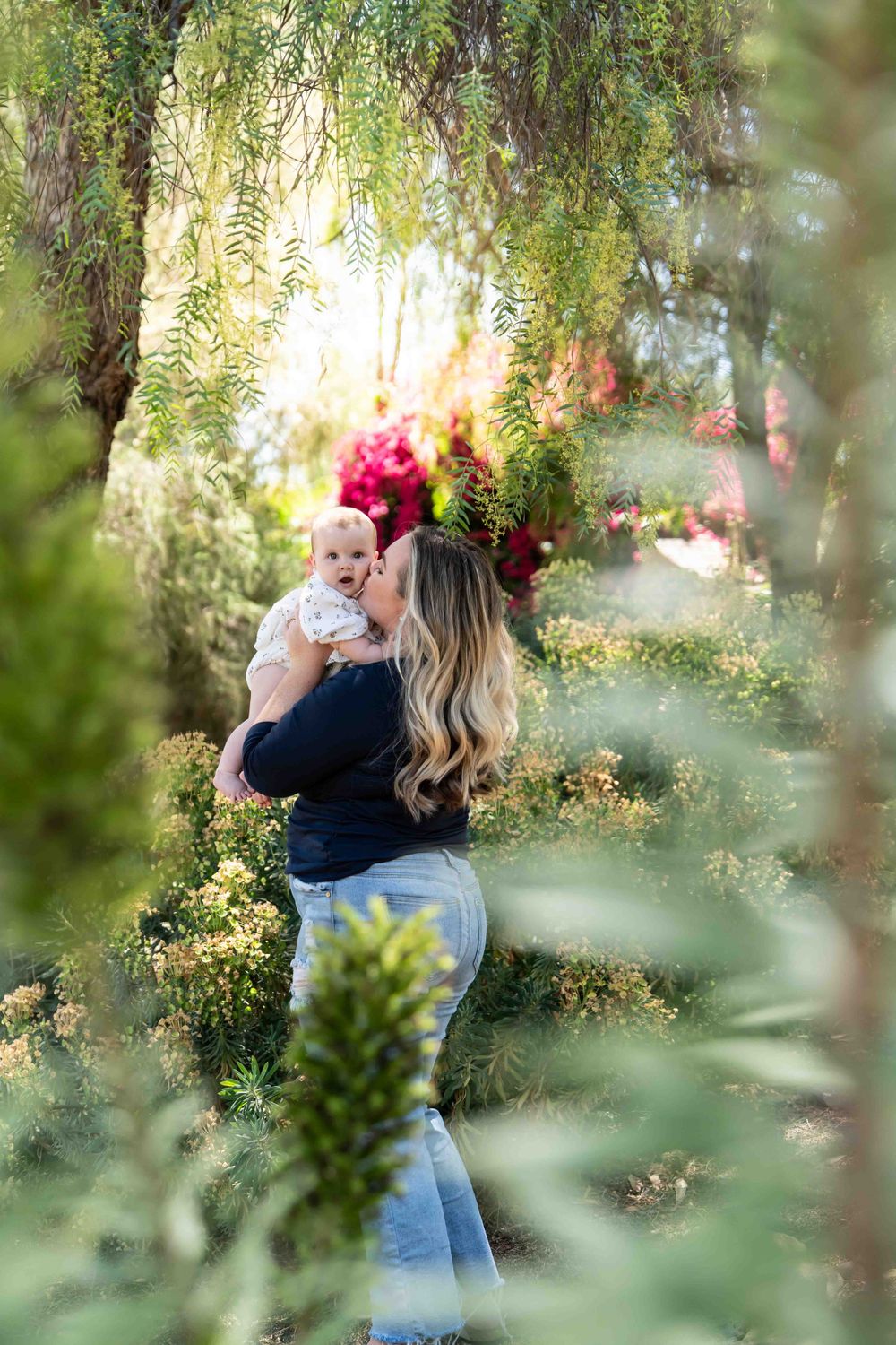 Mother kissing her baby while standing among colorful garden foliage in warm golden sunlight.