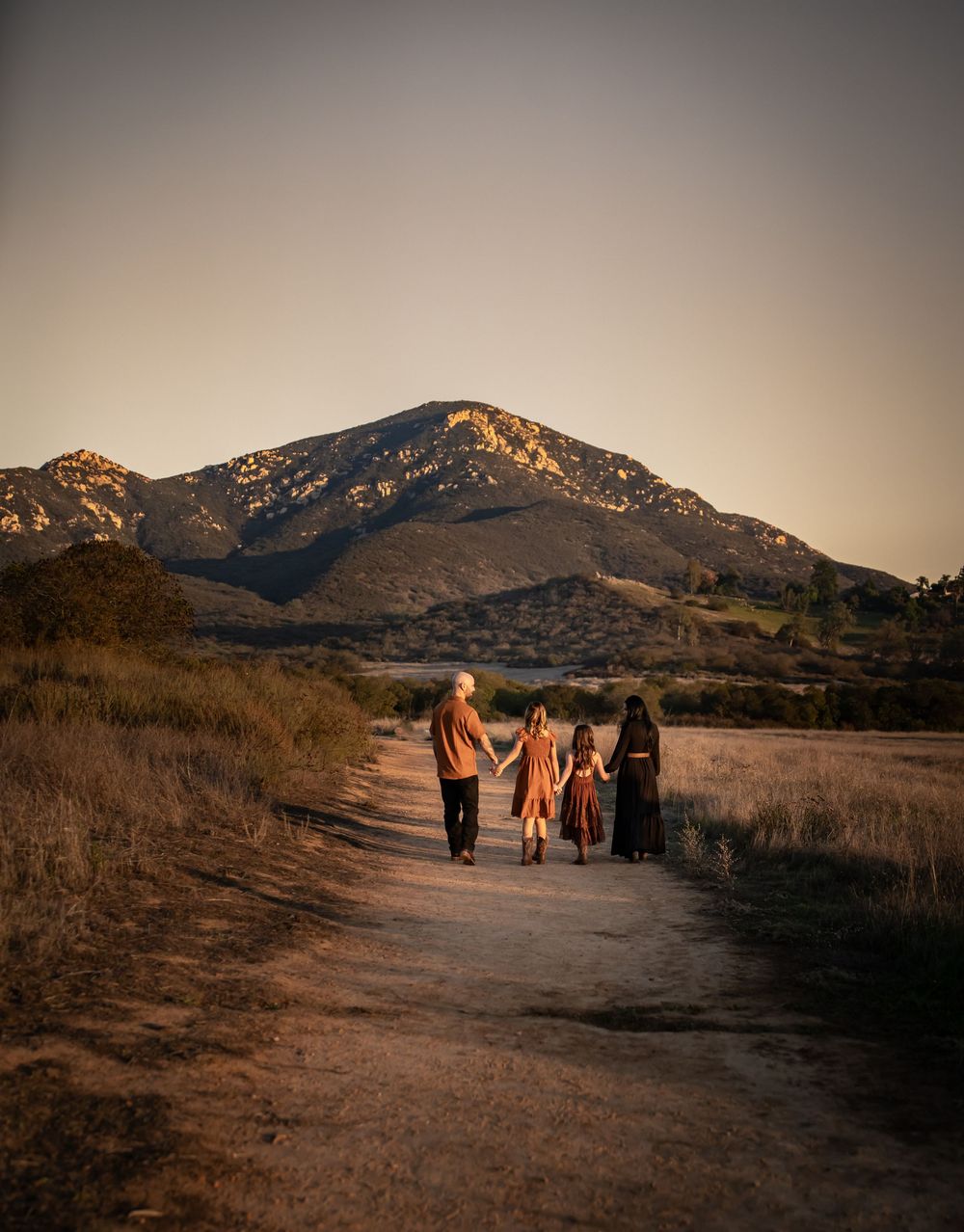 Family of four walking a dirt path toward a sunlit mountain at golden hour in a scenic rural landscape.