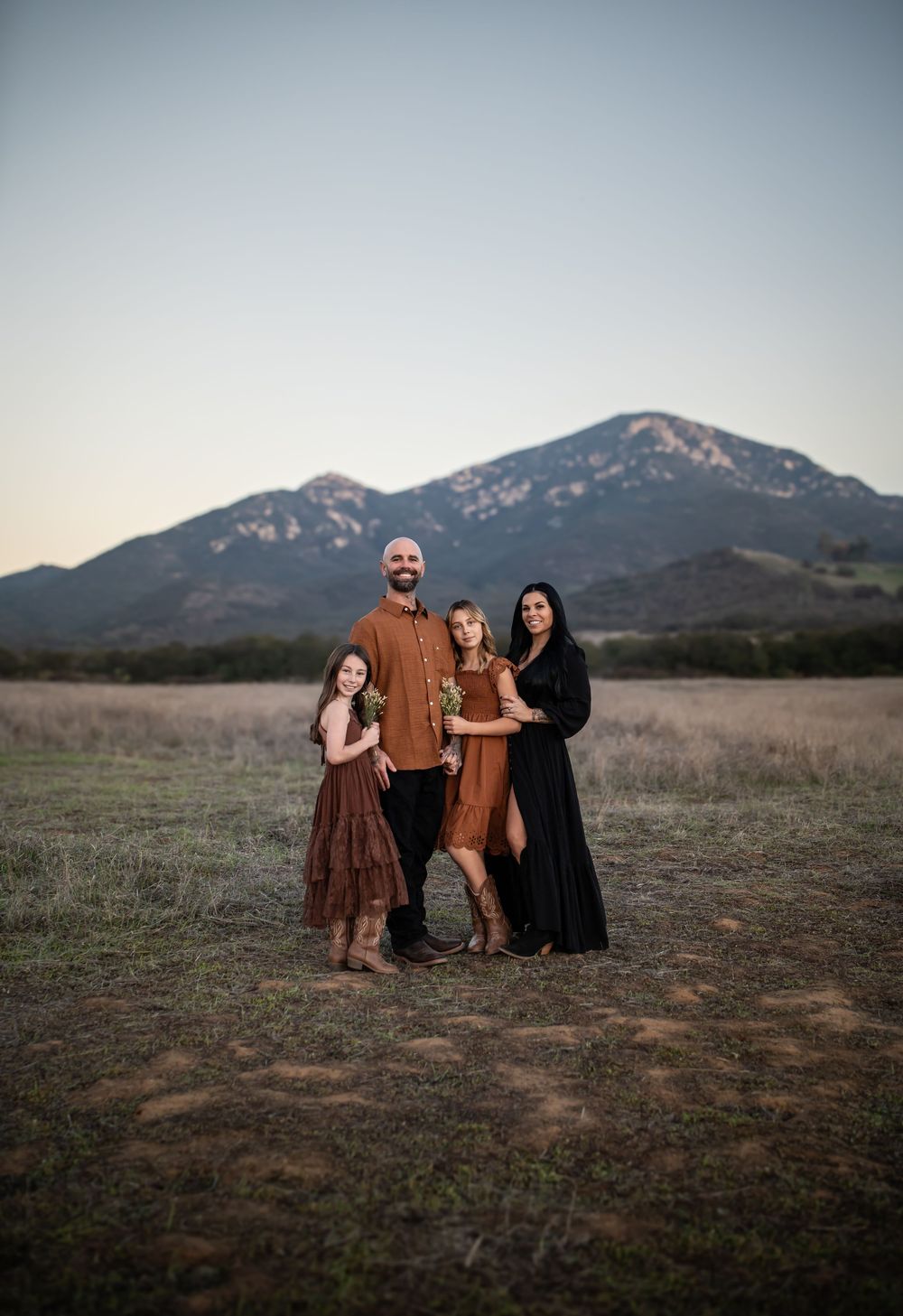Family of four posing in a golden field with mountain backdrop at dusk, wearing earth tones and black.