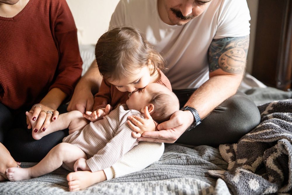 A tattooed father and toddler lovingly kiss a newborn baby while sitting together on a cozy bed. in home session, Ramona