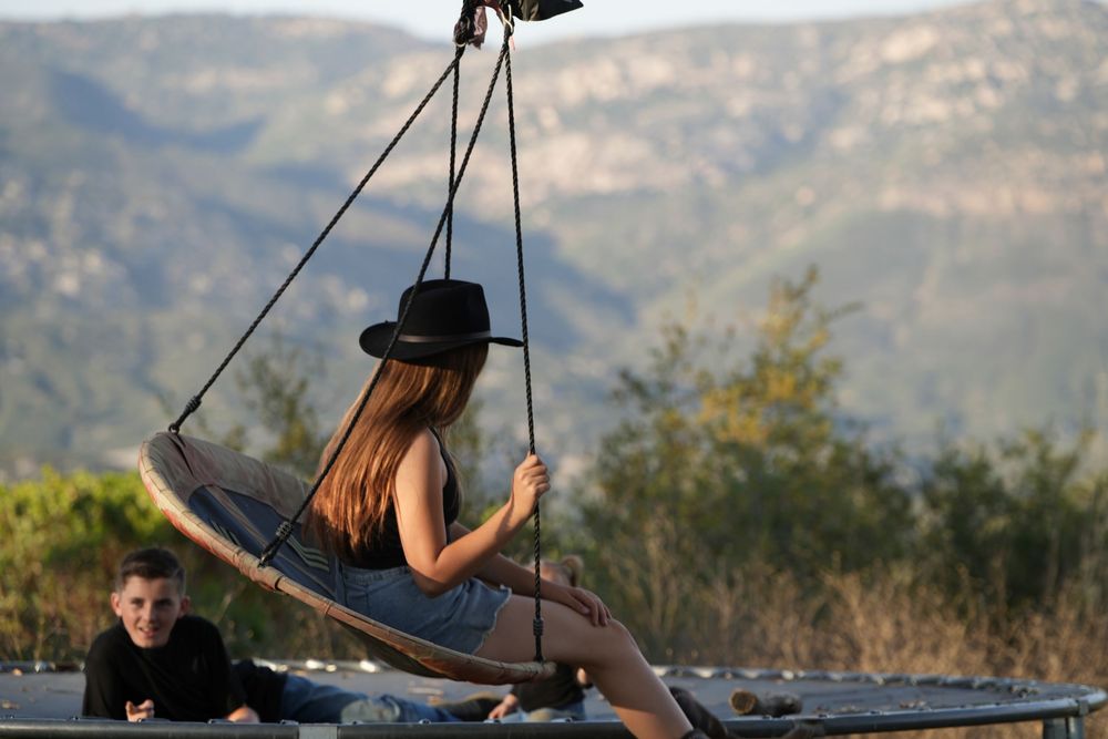 A girl in a black cowboy hat sits on a hanging swing with mountain views in the background.