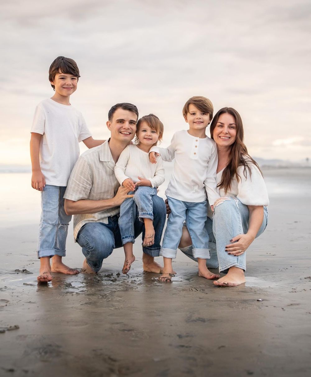 Family of five smiling together on a beach, parents crouching with three young children in white tops and jeans.