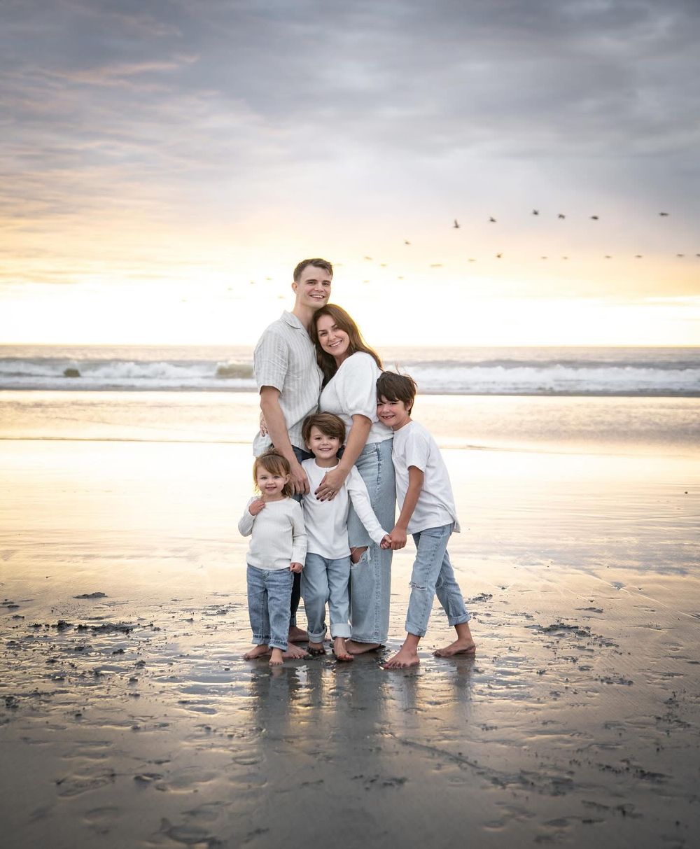 Happy family of five posing at sunset on a beach, parents embracing three children with ocean waves behind them.