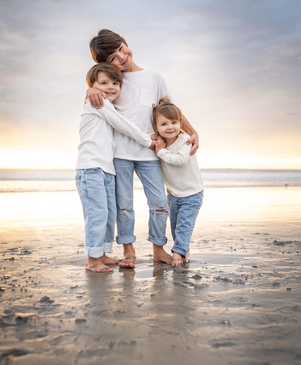 A mother and two children hug on a sandy beach at sunset, all wearing white tops and jeans.