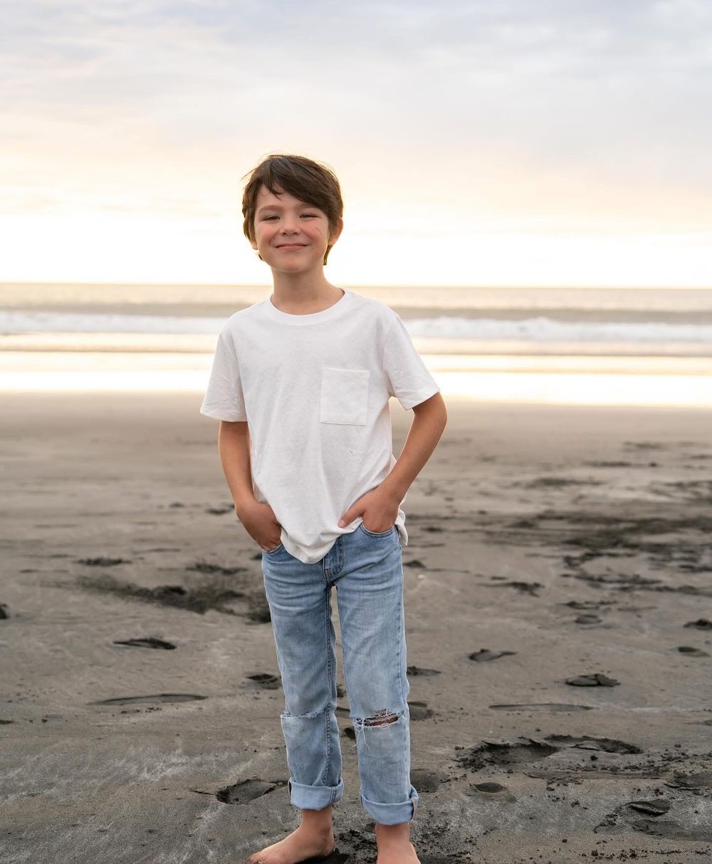 A smiling boy in a white t-shirt and jeans stands barefoot on a sandy beach at sunset.