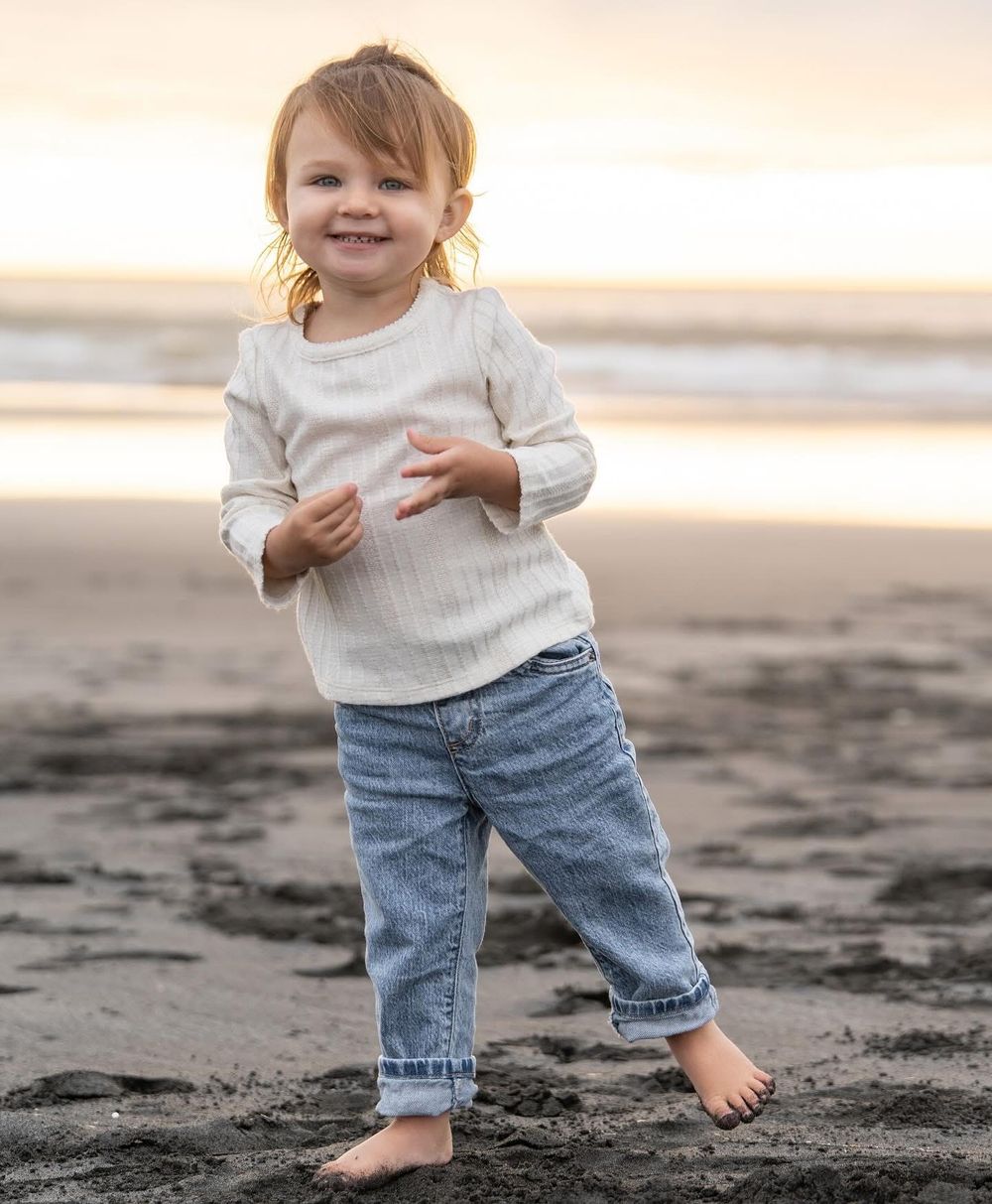 Adorable toddler girl in white top and jeans smiling and standing barefoot on a beach at sunset.