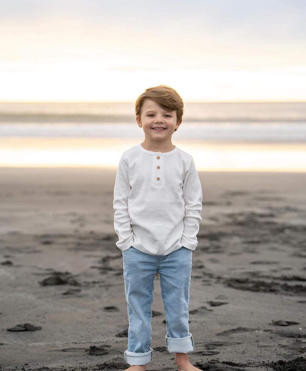 Smiling young boy in white henley shirt and jeans standing on a sandy beach at sunset.