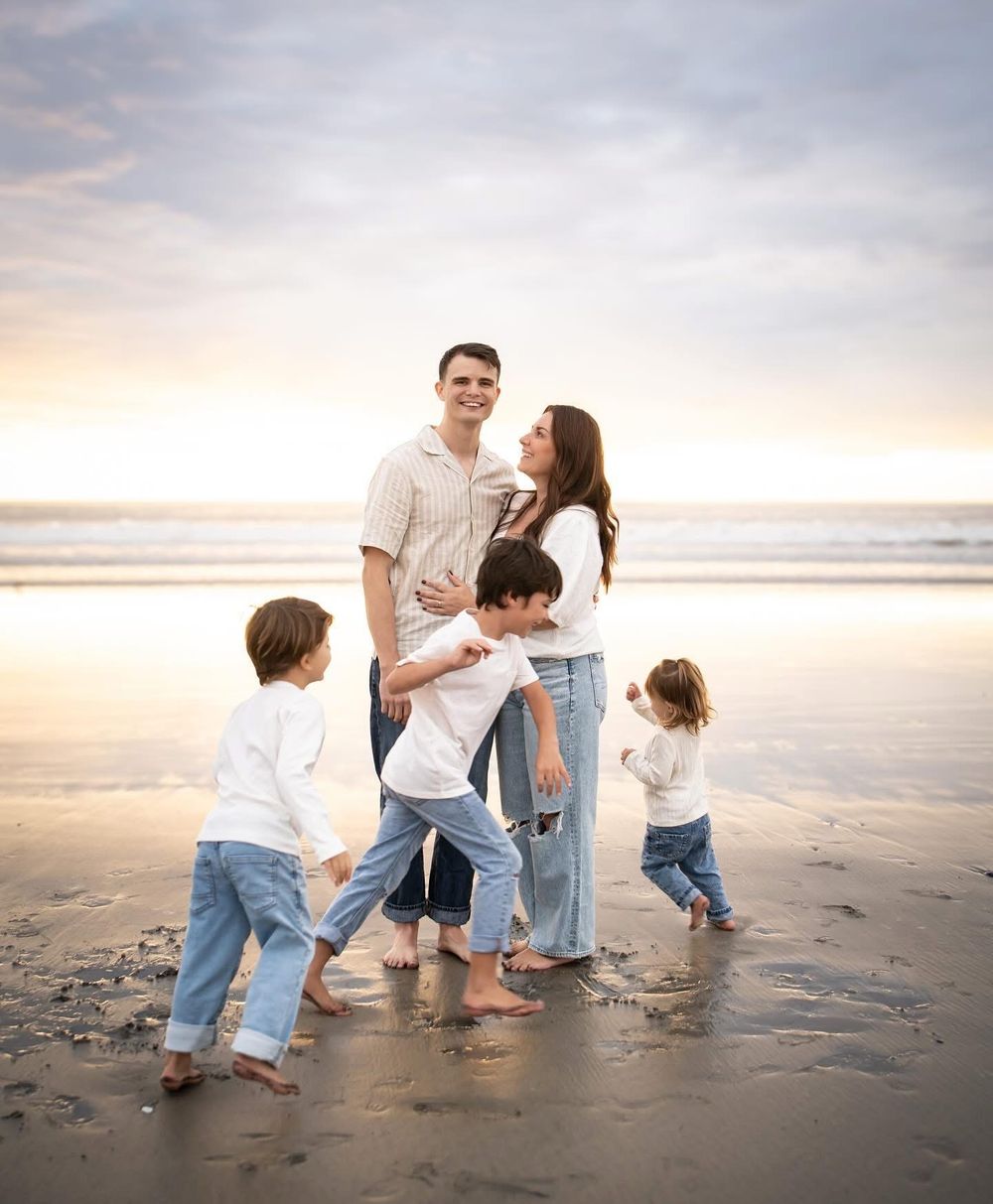 Happy family with three young children posing together on a sandy beach at sunset.