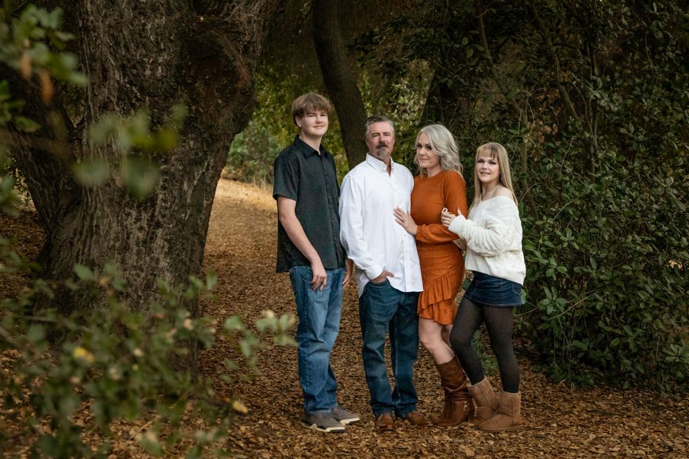 A family of four poses together among tall trees on a leaf-covered path in an autumn forest setting.
