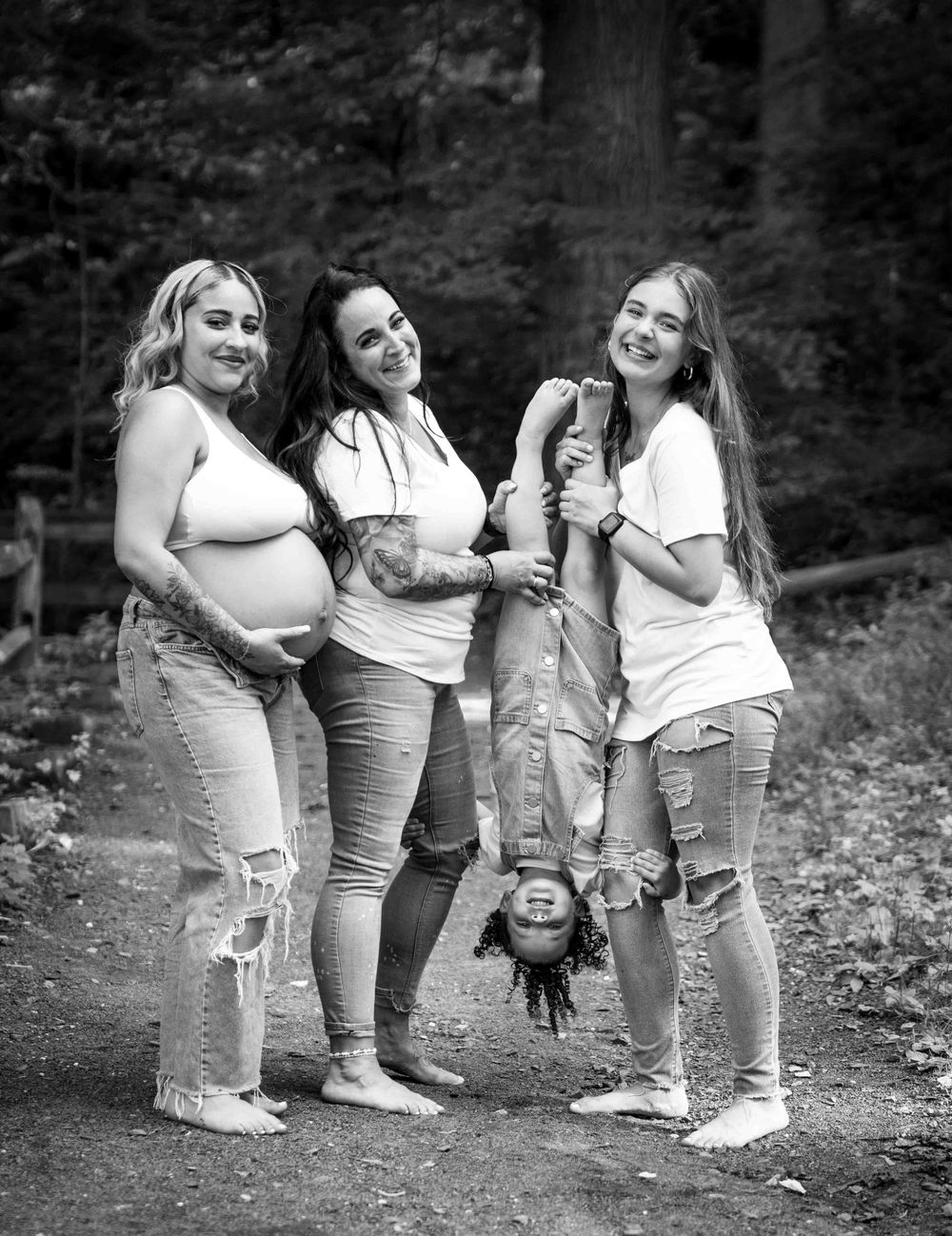 Black and white photo of three women and a child held upside down posing together on a forest path.