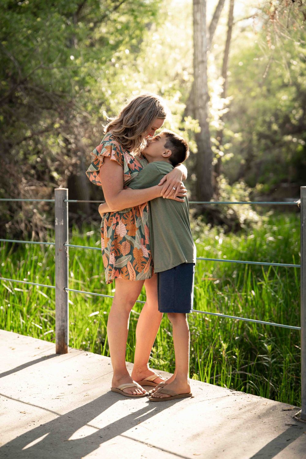 Mother and son sharing a warm hug on a wooden bridge surrounded by lush green nature.