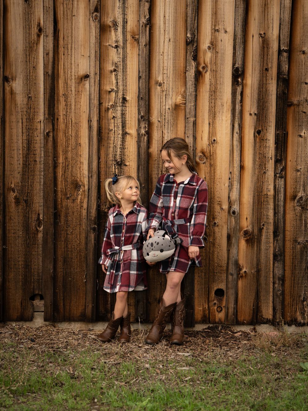 Two girls in matching plaid dresses and brown cowboy boots smile at each other against a rustic wooden fence.