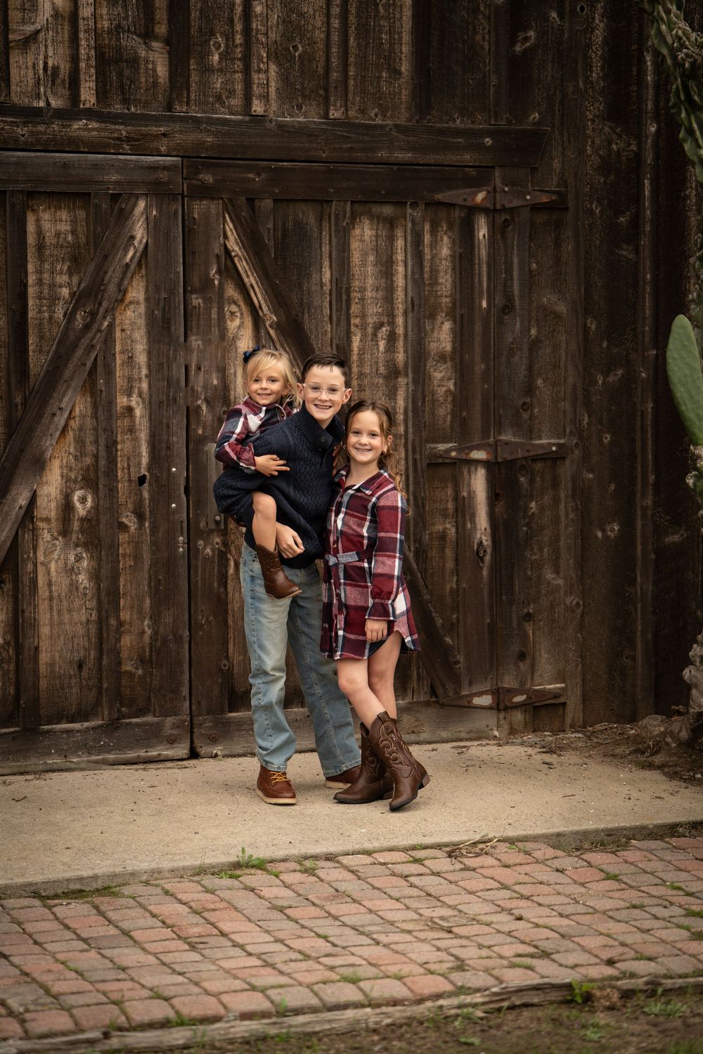Three children pose and smile in front of a rustic wooden barn door, wearing flannel and cowboy boots.