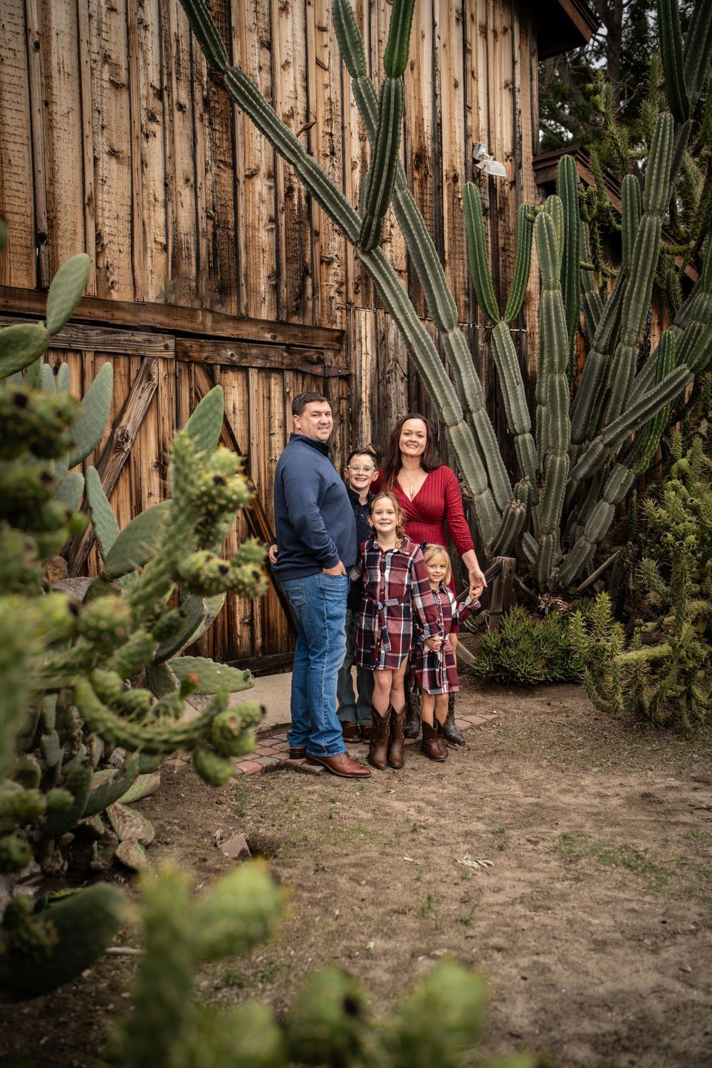 Family of five posing together near tall cacti and a rustic wooden barn wall outdoors.