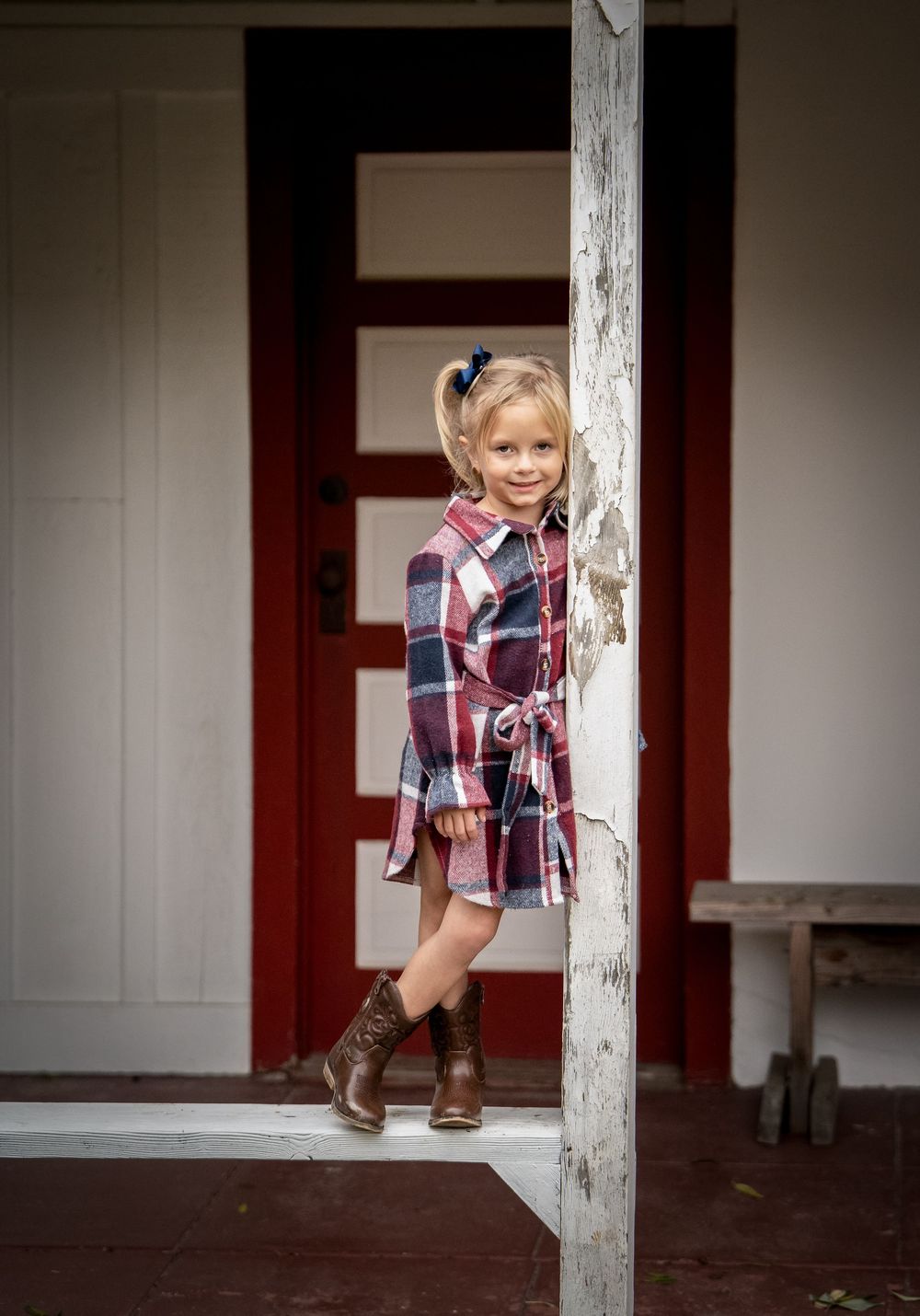 Young girl in plaid dress and brown cowboy boots poses by a white porch post near a red door.