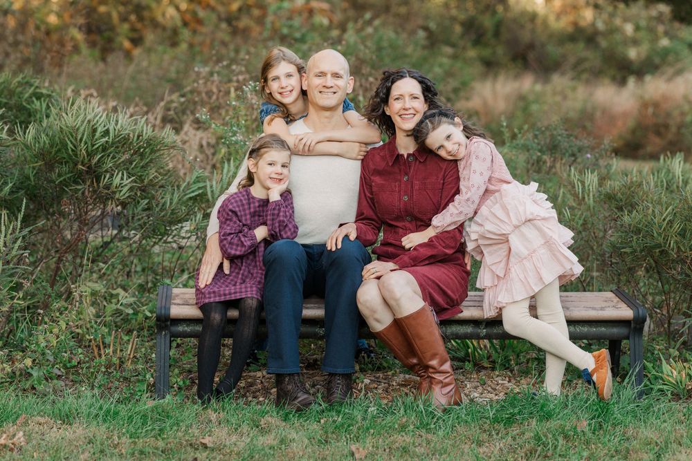 A family sits together on a wooden bench in a grassy outdoor setting during golden hour.
